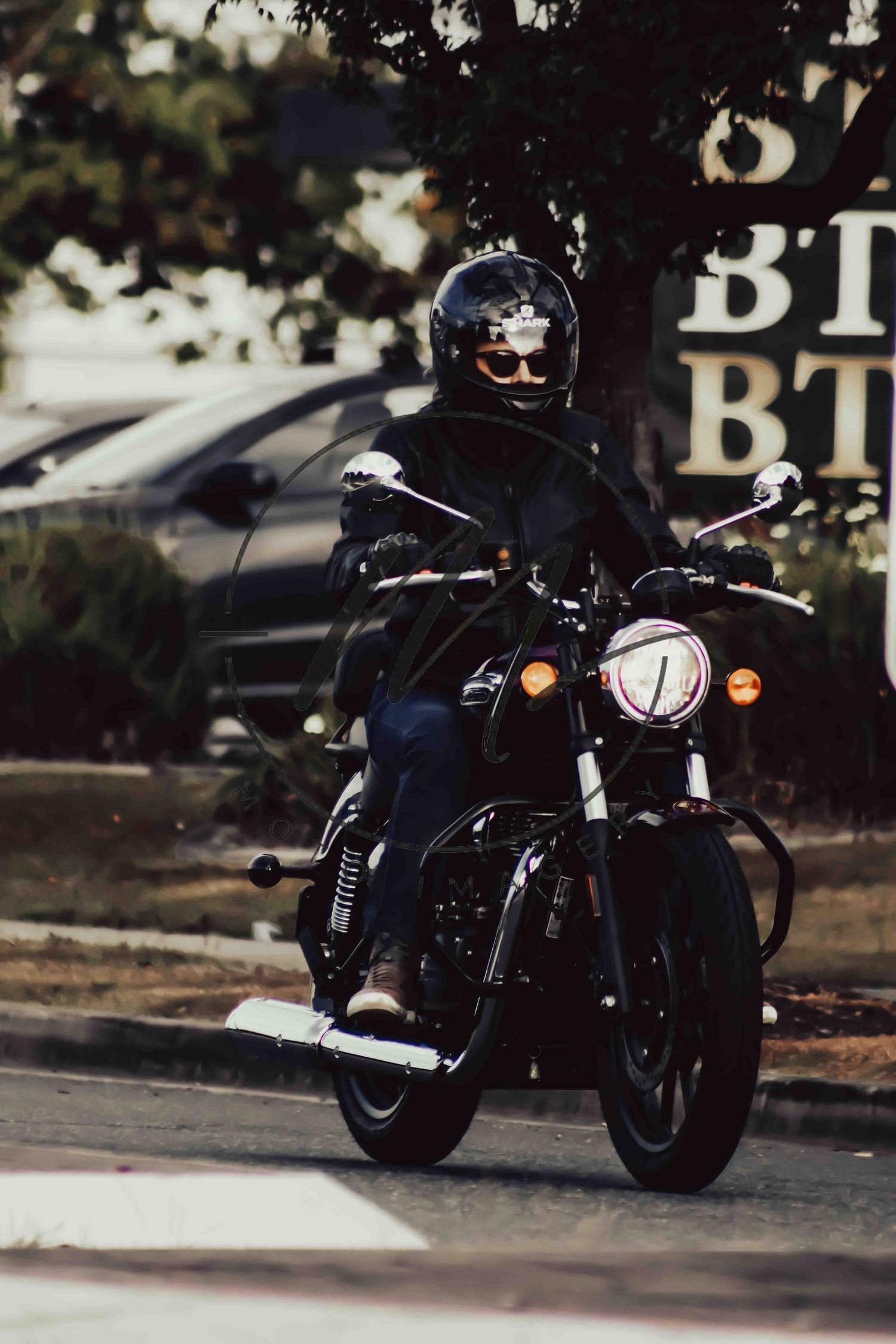 A motorcyclist in black helmet and jacket rides a black cruiser motorcycle on a street near parked cars.