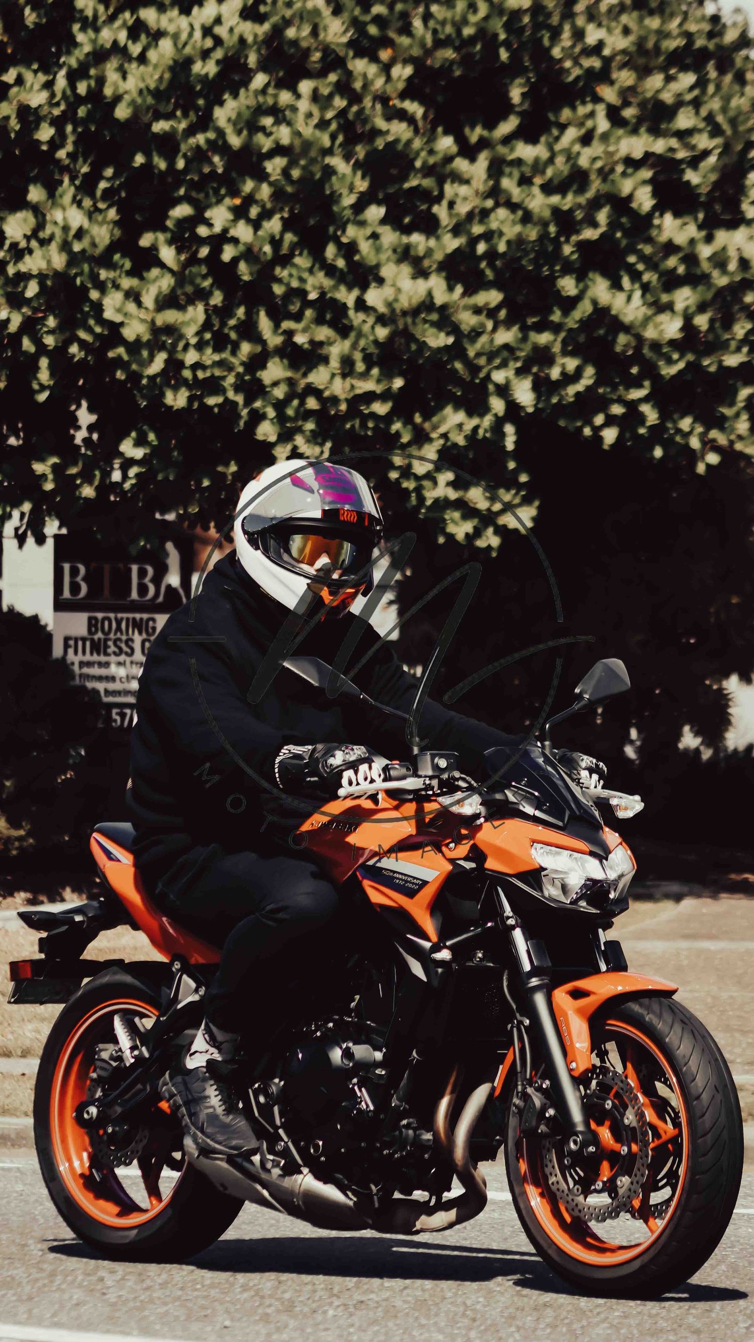 A rider in a white helmet and goggles on an orange sport motorcycle parked on a sunny street near a boxing fitness center.