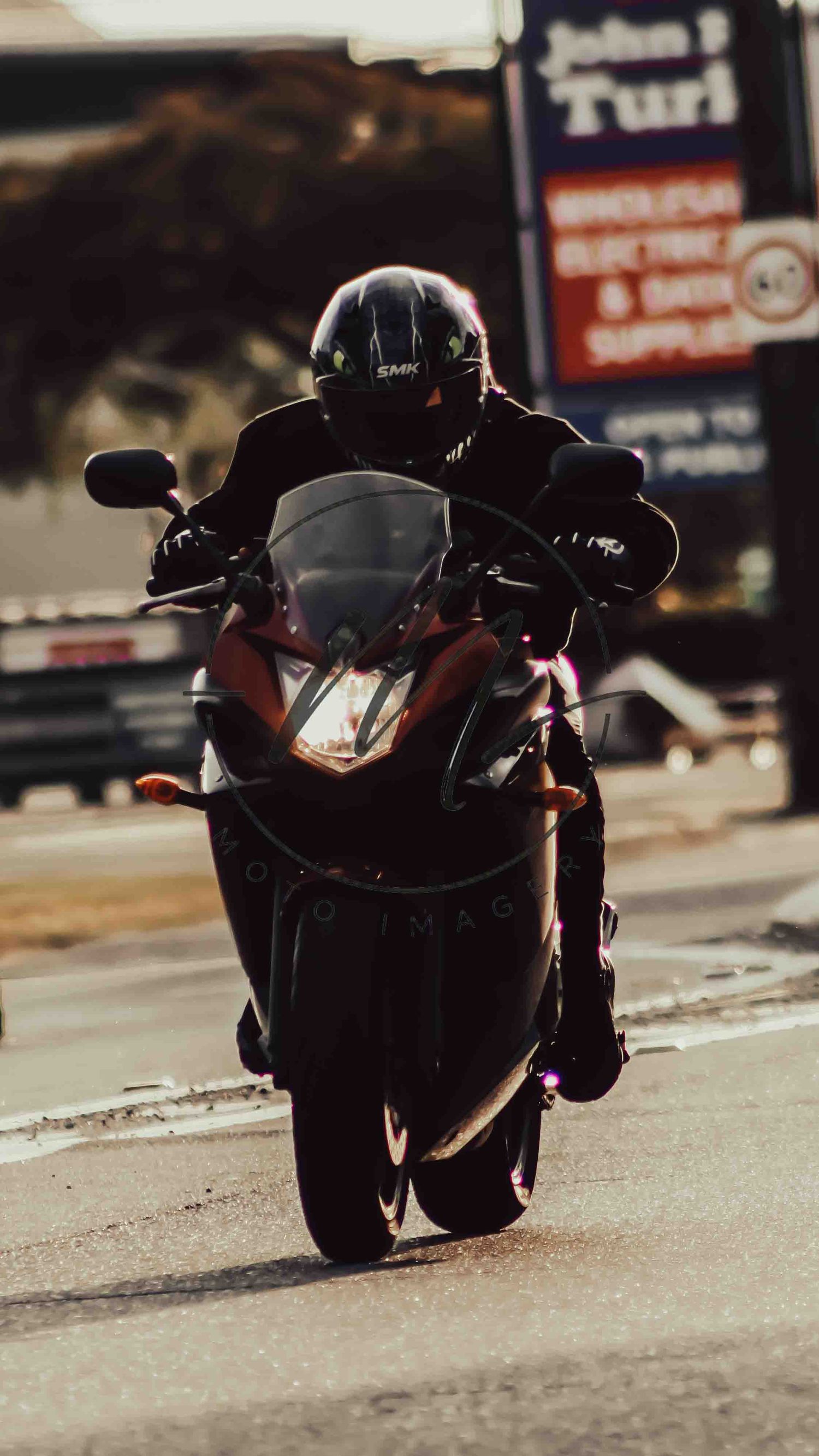 A motorcyclist in black gear and helmet rides a red sport bike on a city street near an electric supply store.