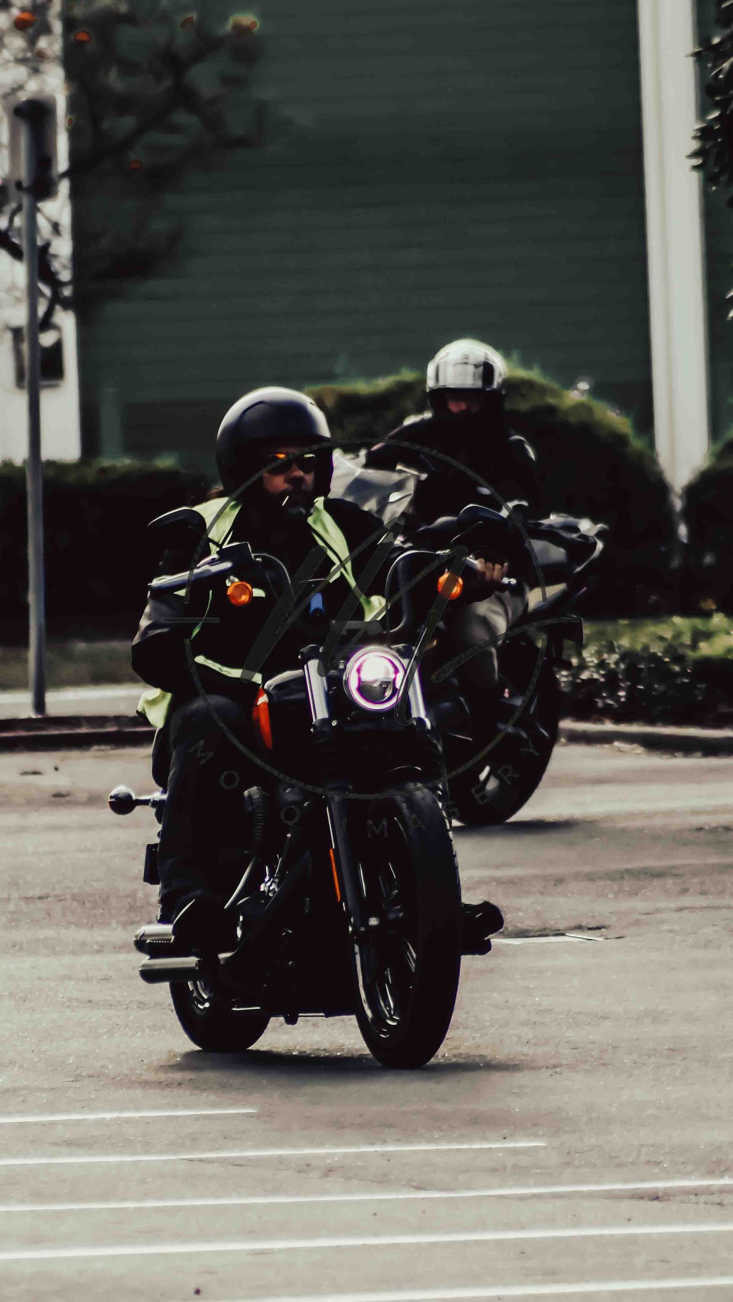 Two motorcyclists in helmets and gear ride black motorcycles on a city street.