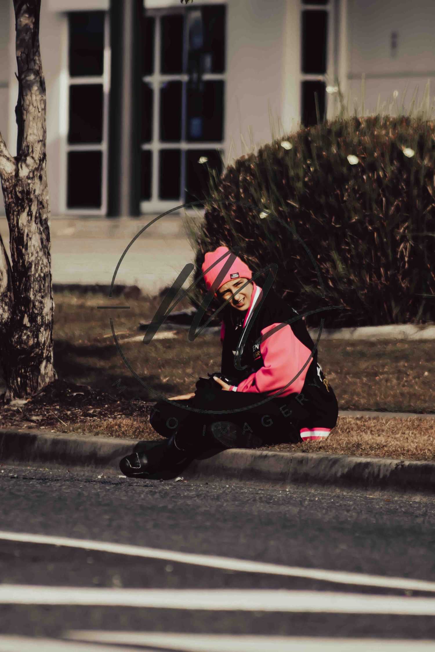 Person in pink hoodie and hat sitting on curb near road, looking at phone outdoors.