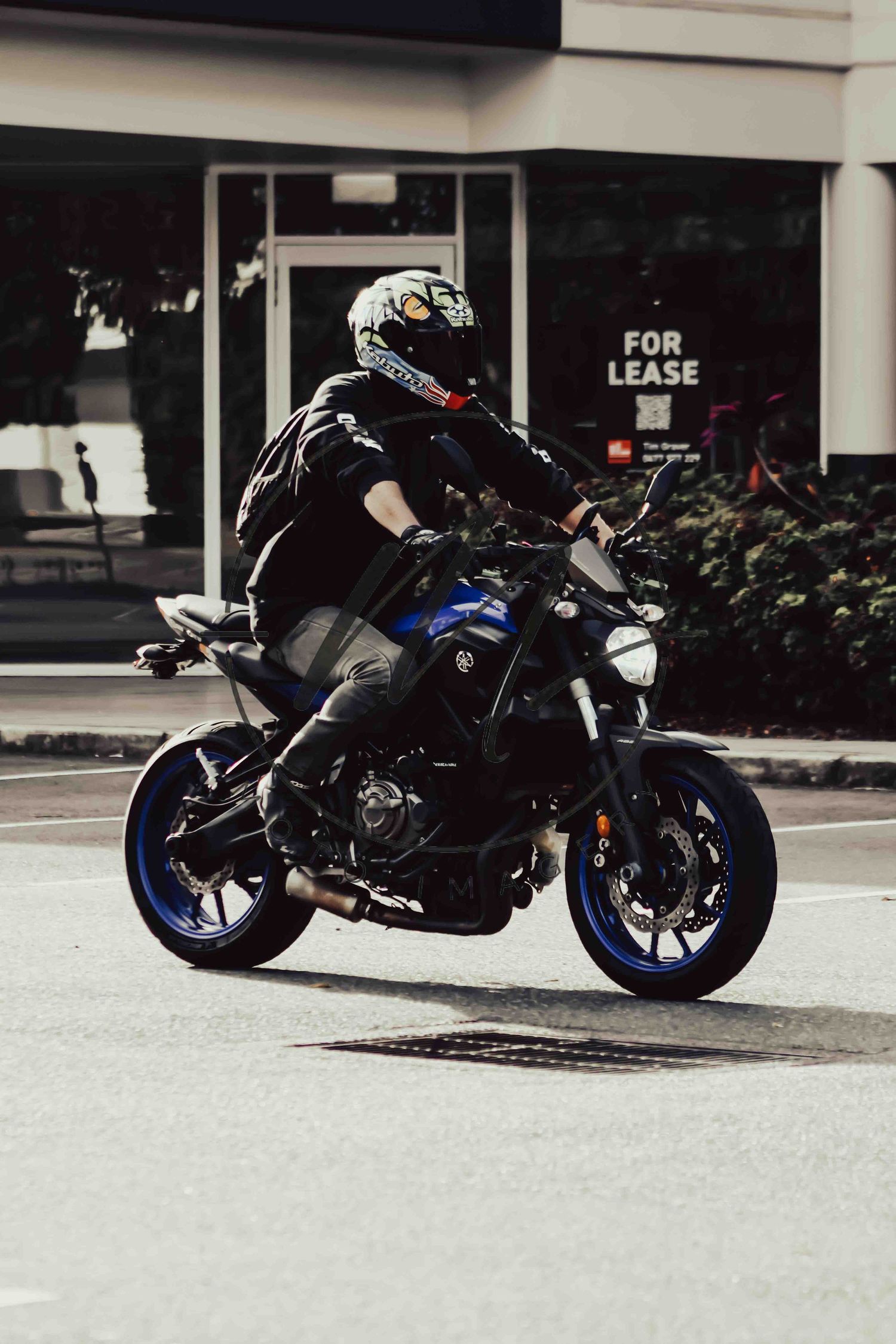 Motorcyclist in black jacket and helmet riding a blue Yamaha MT sport bike on an urban street past a for lease sign.