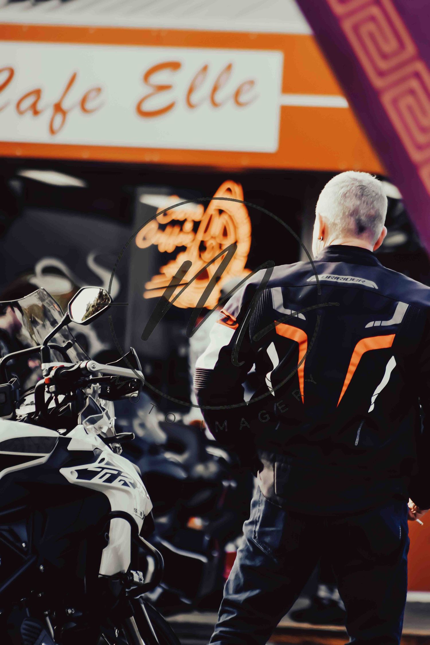 Gray-haired motorcyclist in black and orange jacket stands beside his bike outside Cafe Elle.
