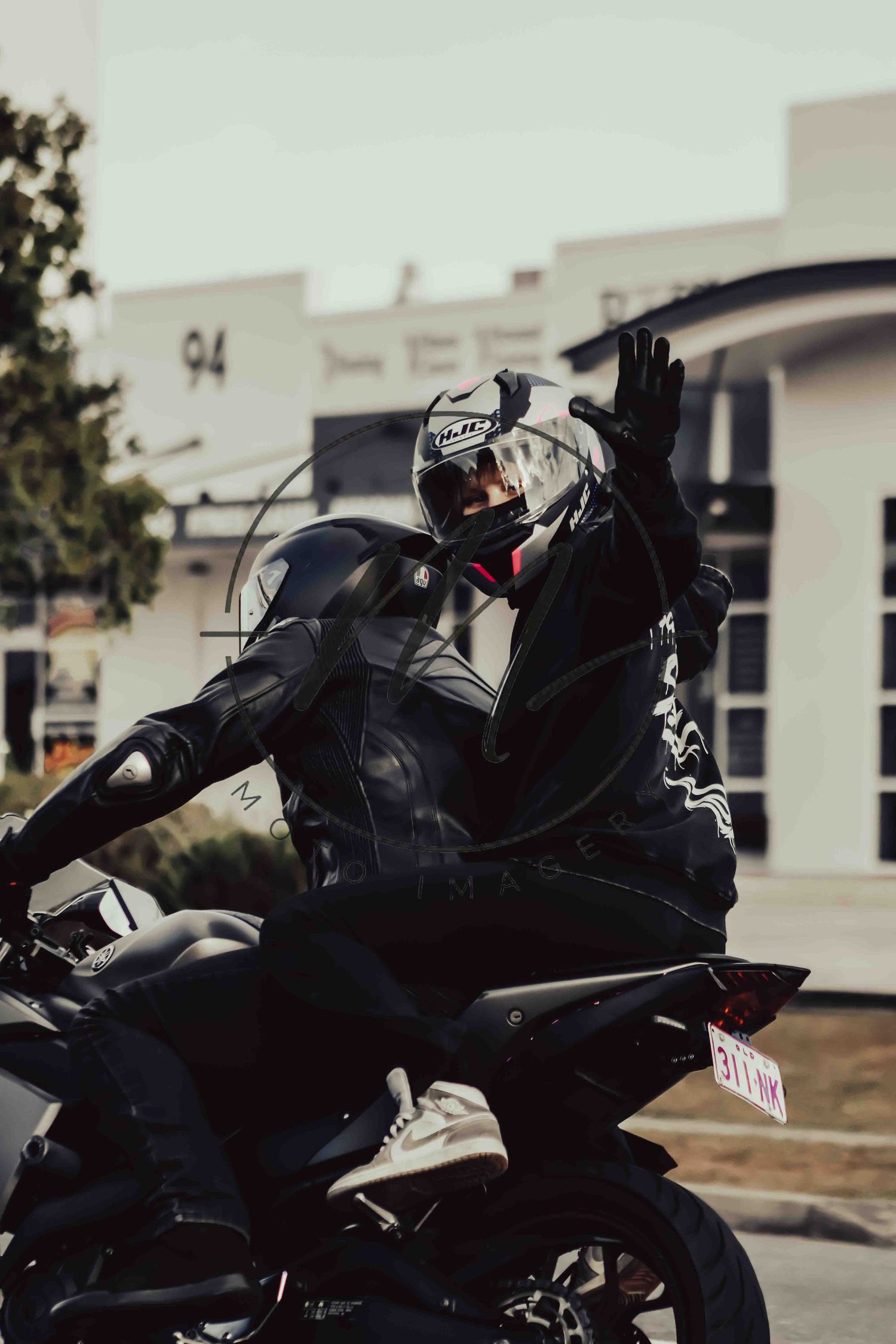 A motorcyclist in black leather gear and white helmet waves while sitting on a sport bike outside a building.