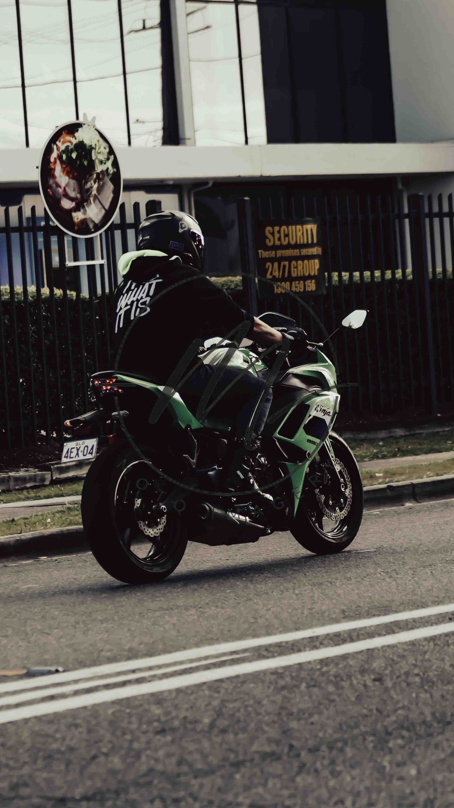 Rider in black jacket on a green and black sport motorcycle parked on a city street near a security sign.