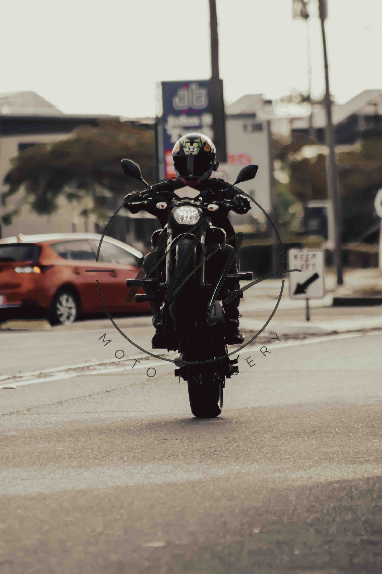 Motorcyclist performing a wheelie on a dark motorcycle at an urban intersection, wearing a helmet, with cars and buildings in background.