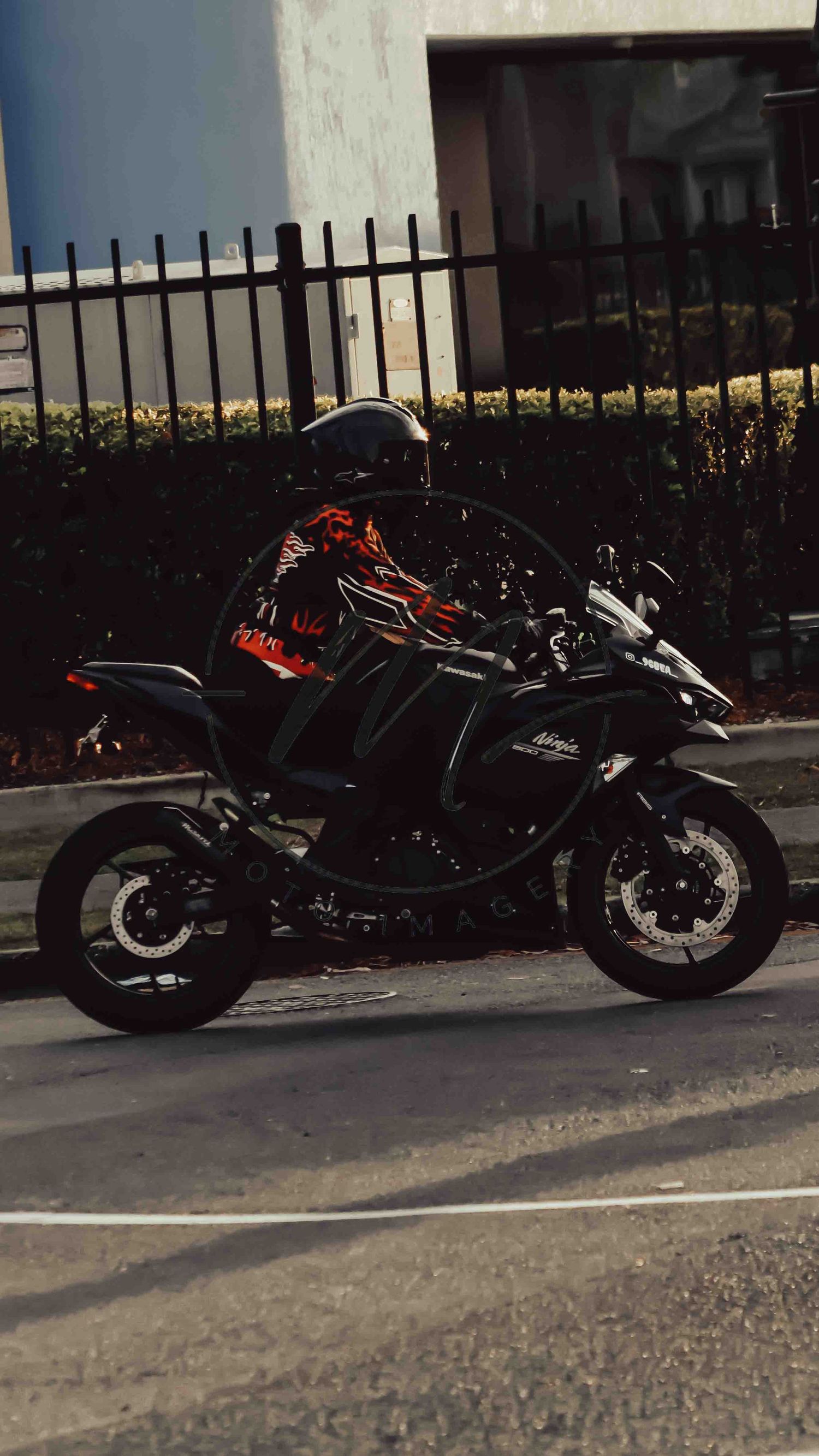 A rider in an orange and black jacket sits on a matte black Kawasaki Ninja motorcycle on a city street.