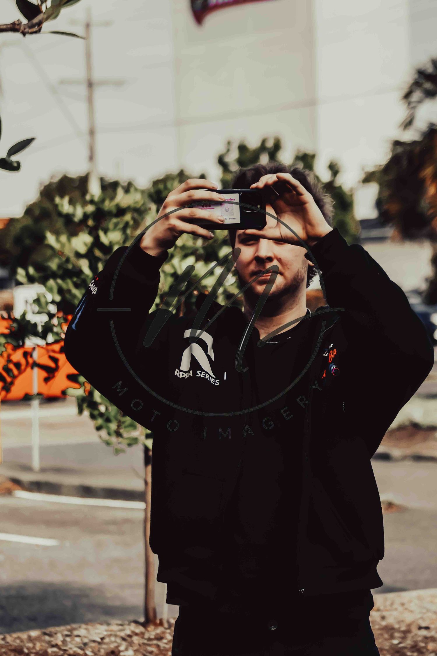 Man in black hoodie holds smartphone up to take a photo outdoors near trees and a street.