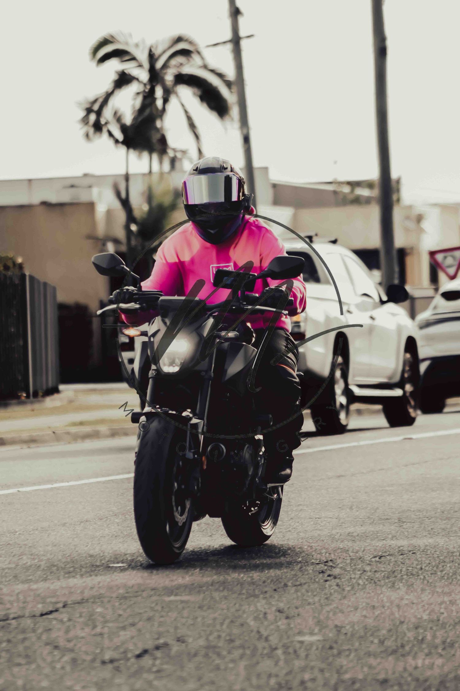 Motorcyclist in pink shirt and black helmet riding a dark sport motorcycle on a urban street with palm trees.