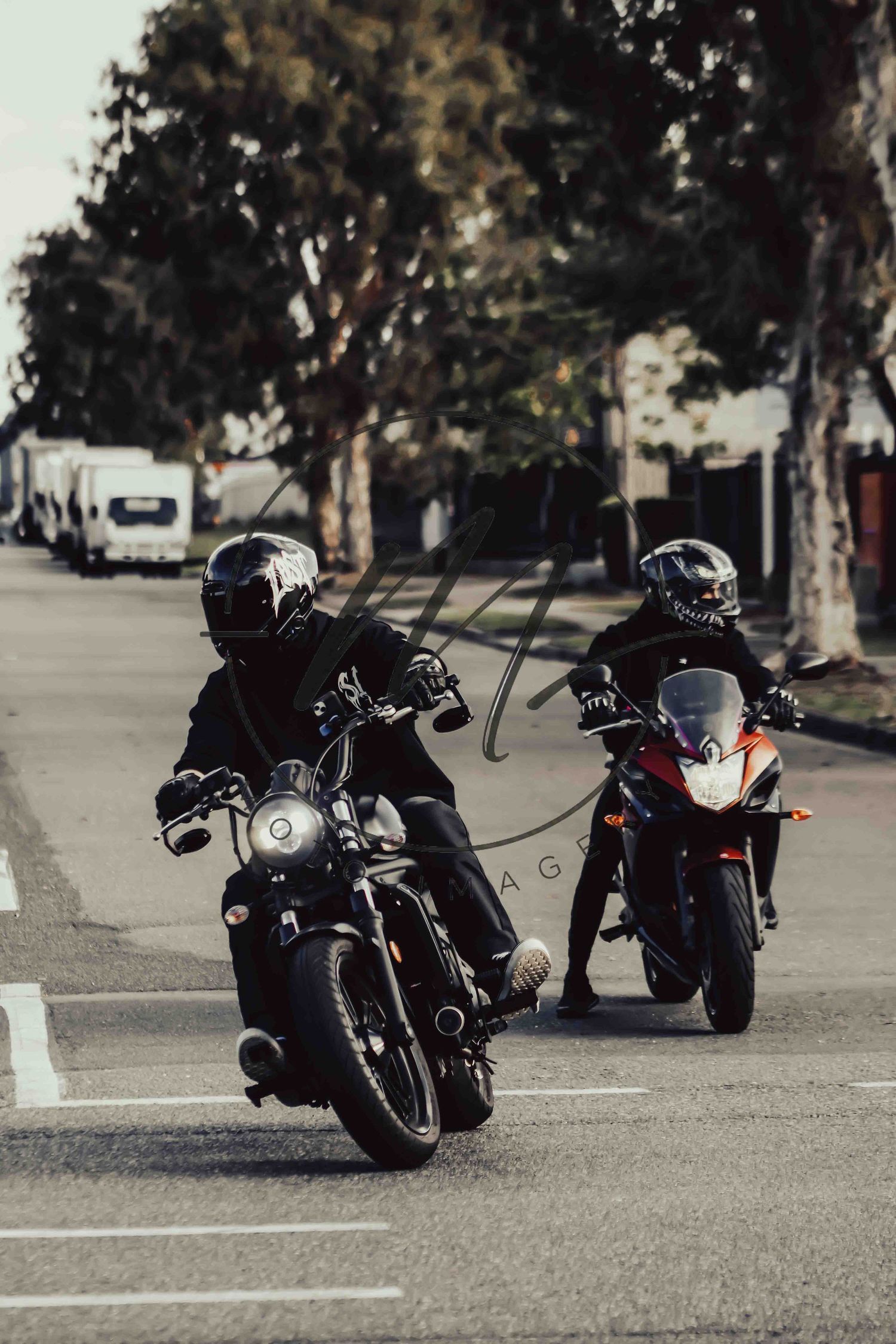 Two motorcyclists in black gear and helmets riding on a tree-lined street.