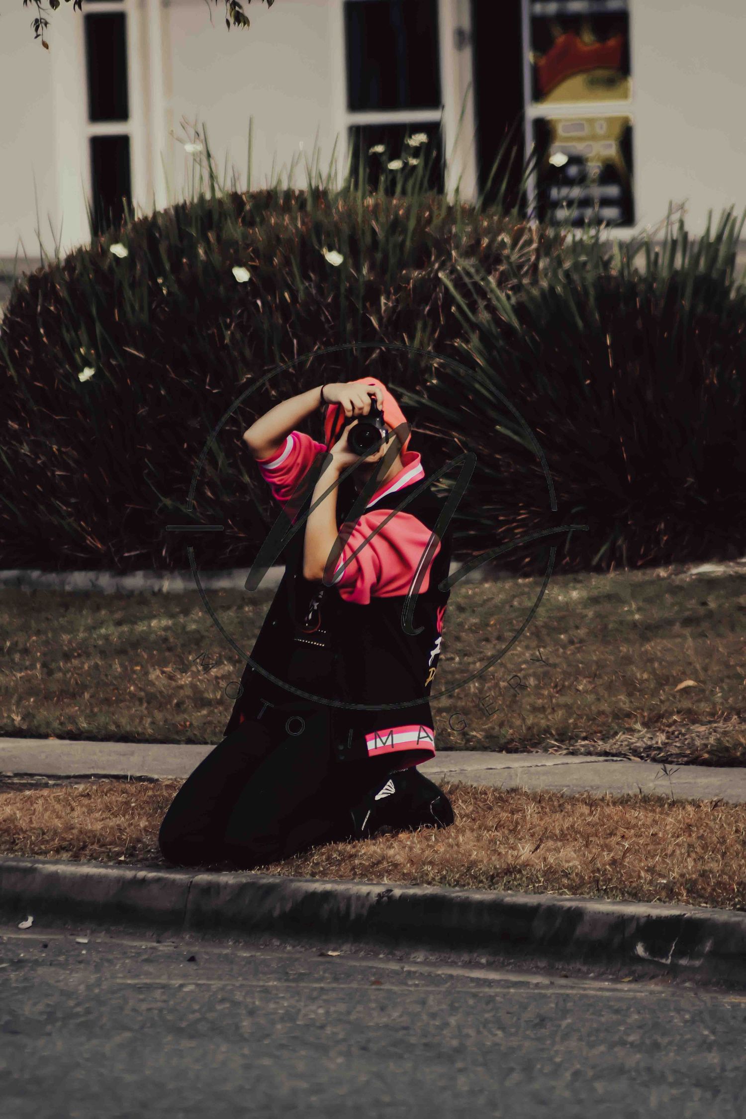 Person kneeling on grass photographing with a camera, wearing black and red outfit, with green shrubs and building in background.