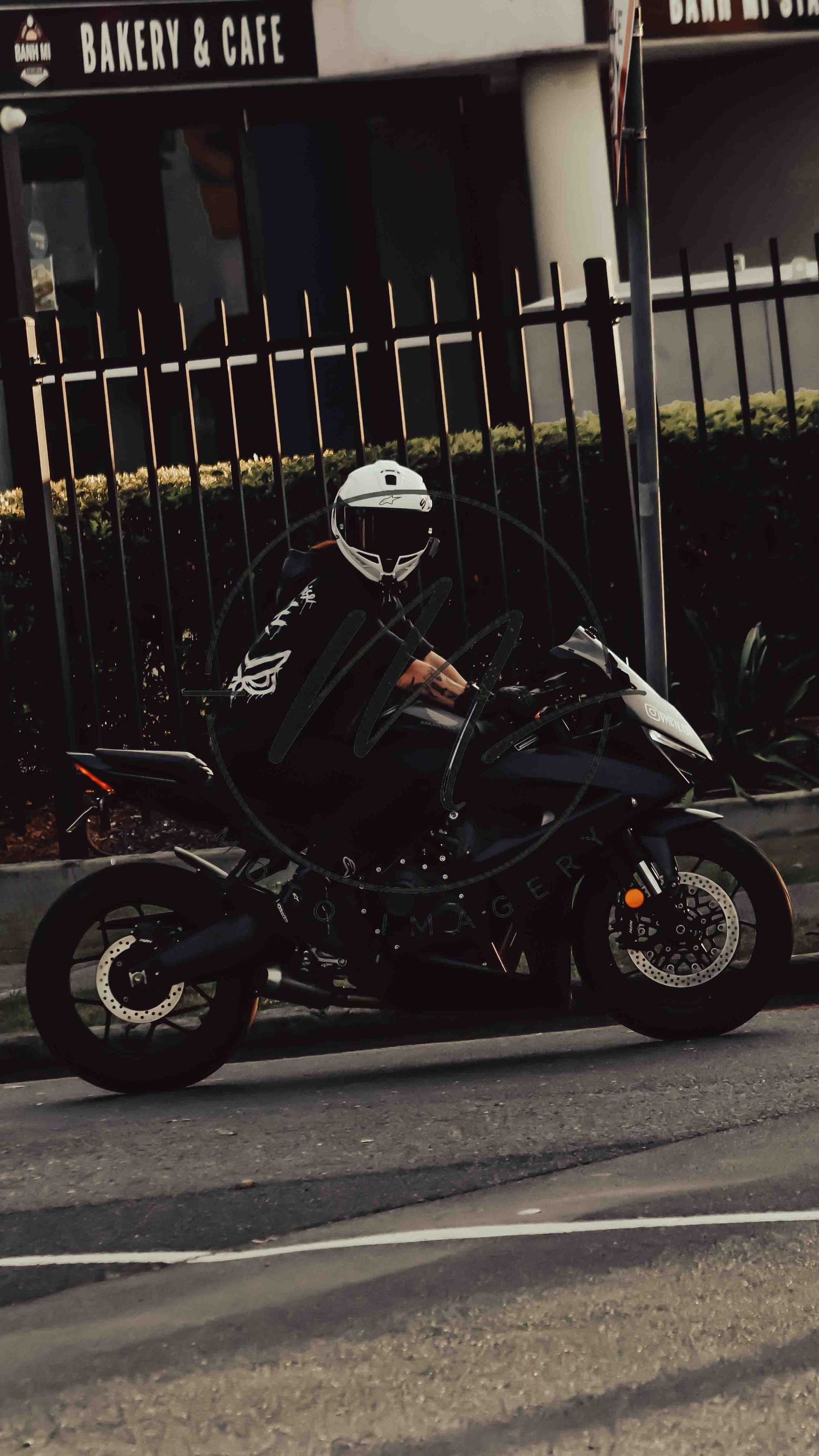 A motorcyclist in a white helmet and black jacket rides a black sport bike near a bakery and cafe.