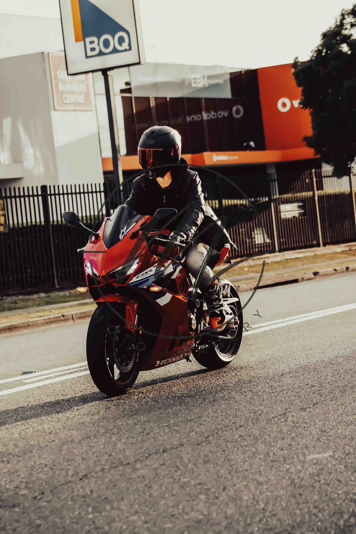 Motorcyclist in black gear riding red sport bike on urban street near BOQ bank branch.