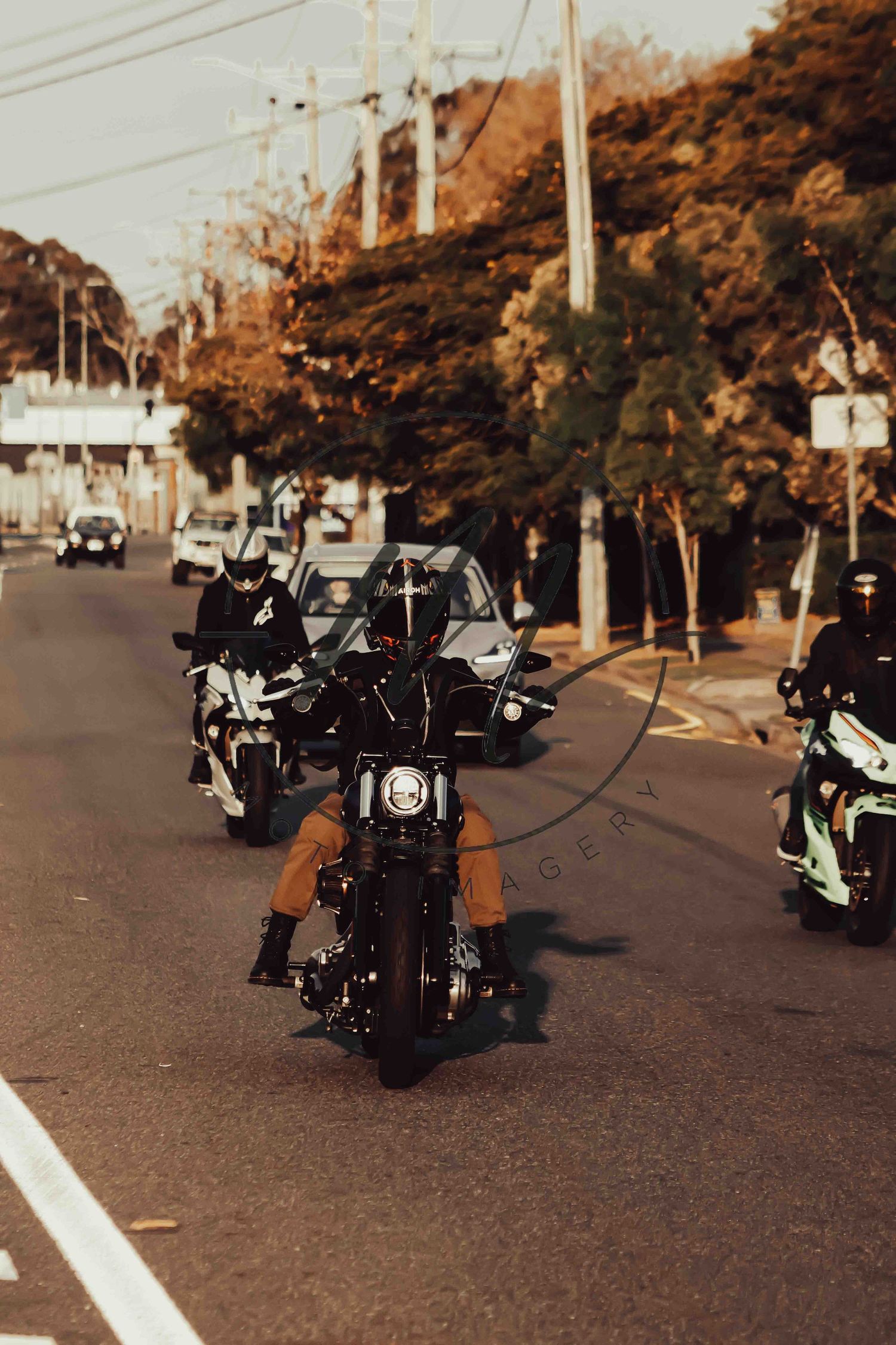 Group of motorcyclists riding in formation down a tree-lined street on classic black motorcycles.