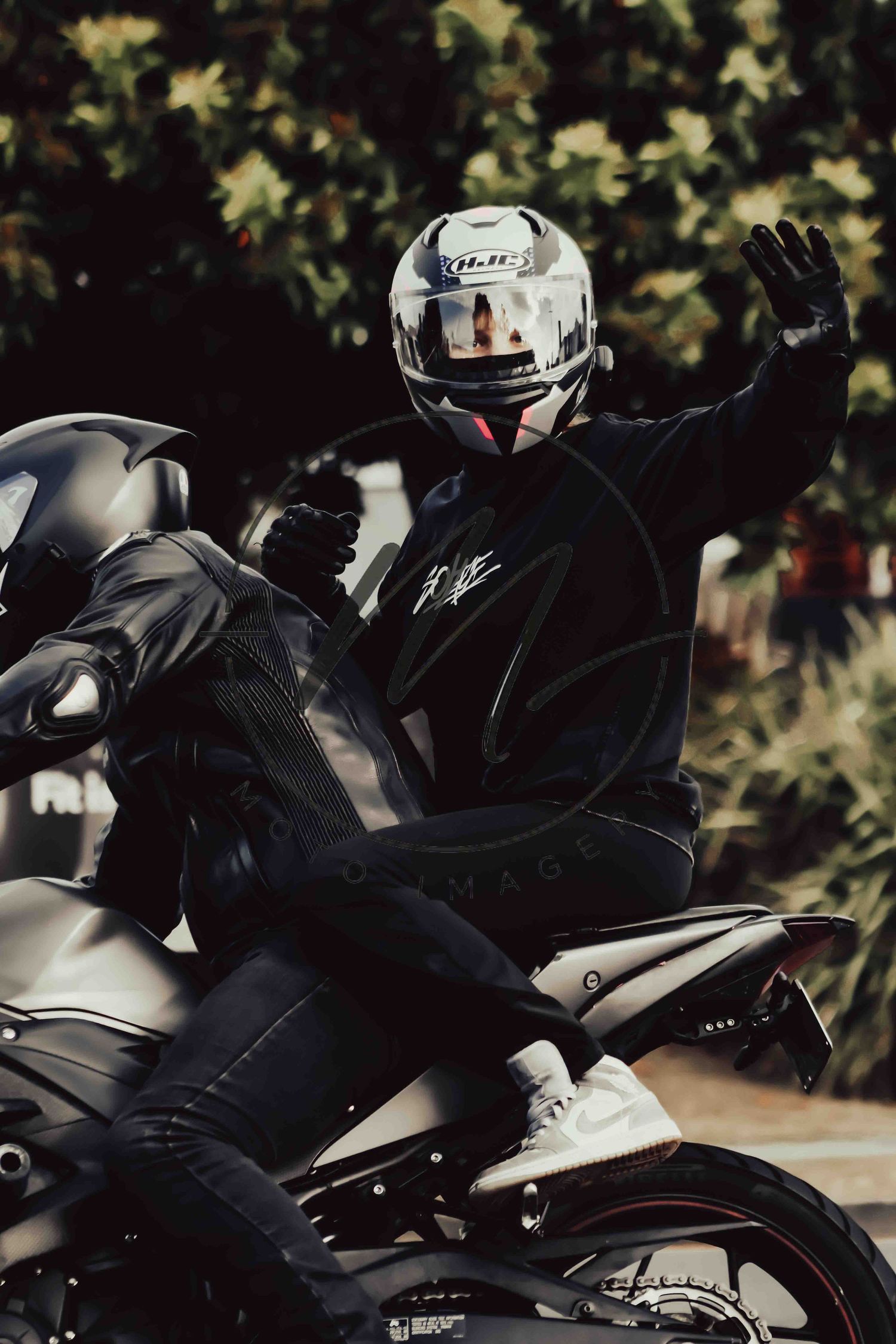 Motorcyclist in black leather jacket and white HJC helmet waving while seated on a sport motorcycle outdoors.