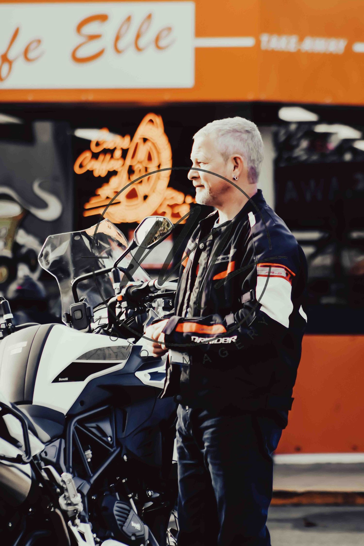 Older man with white hair in motorcycle jacket standing beside a touring bike outside a cafe.