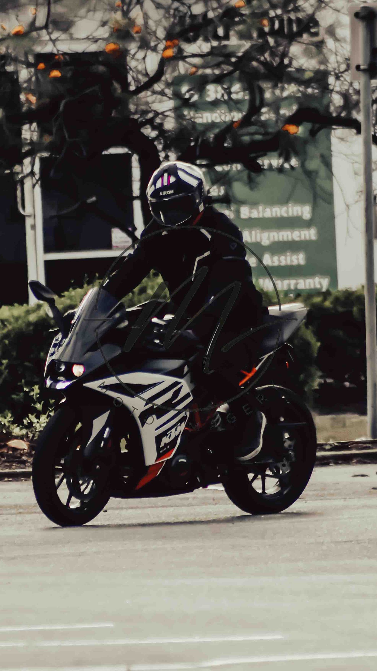 A rider in black gear and helmet navigates a KTM RC sport motorcycle on a city street near an auto service shop.