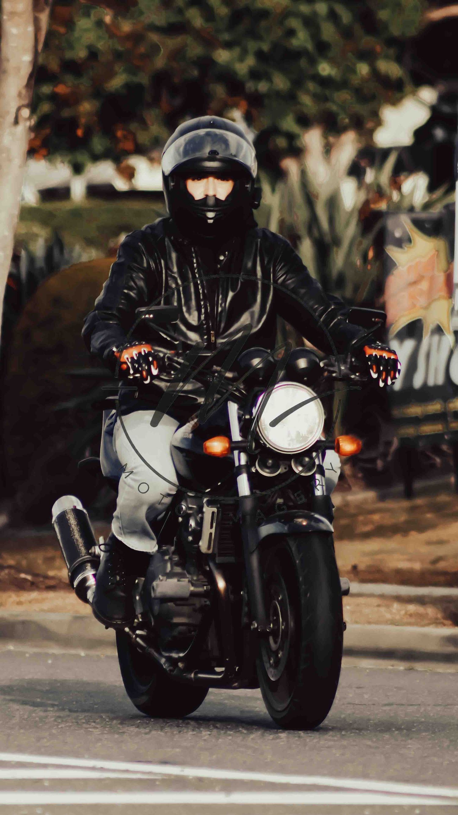 Motorcyclist in black leather jacket and helmet riding a classic motorcycle on a street.