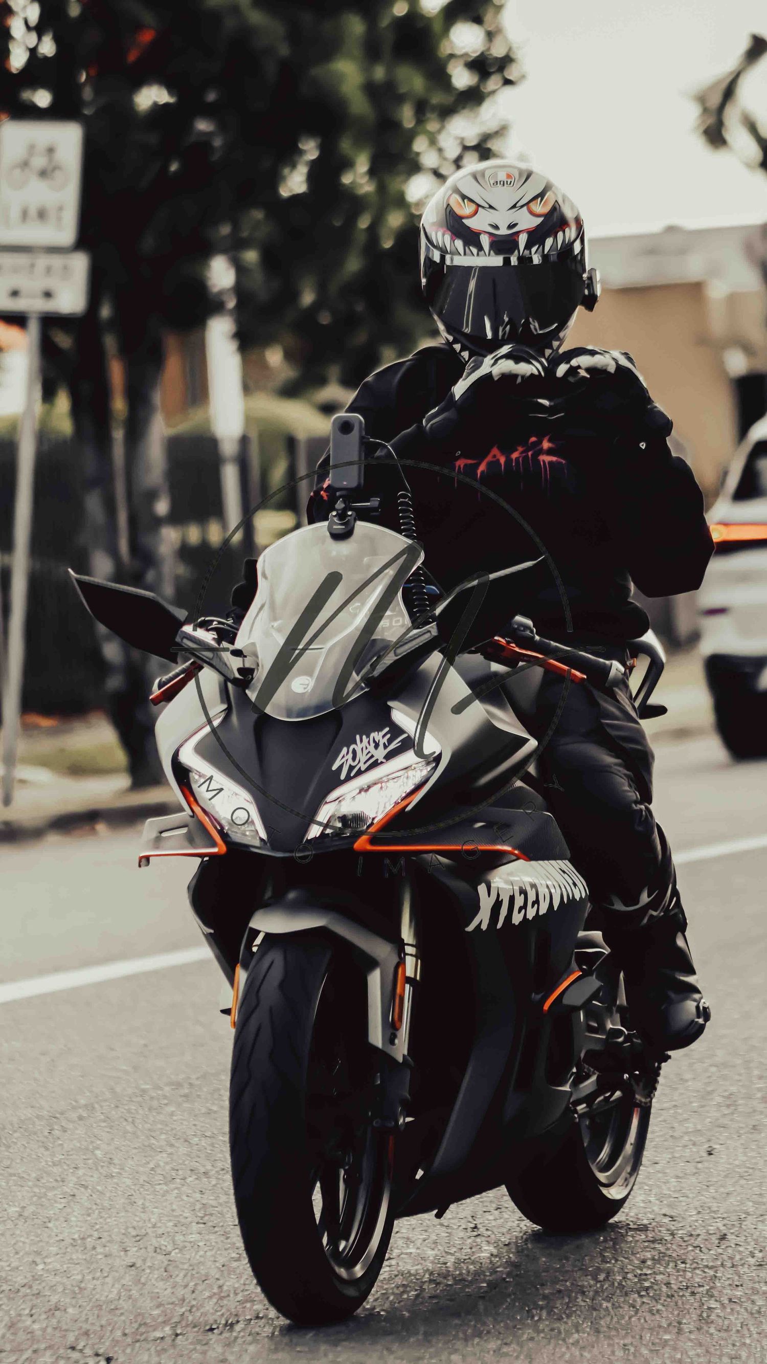 Motorcyclist in black gear and menacing helmet rides a sport bike with orange accents on a city street.