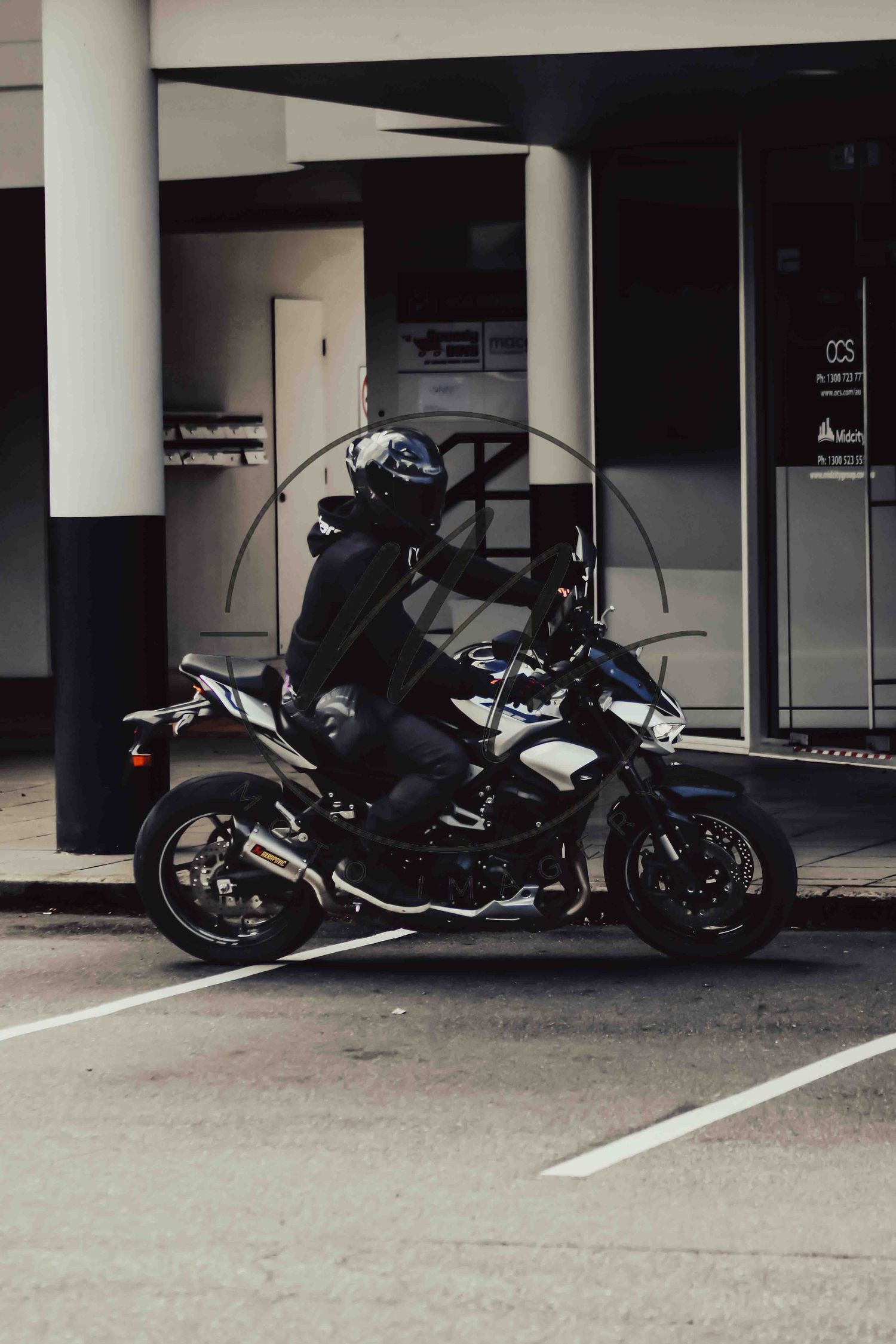 A motorcyclist in black gear and helmet rides a dark sport motorcycle near a modern building.