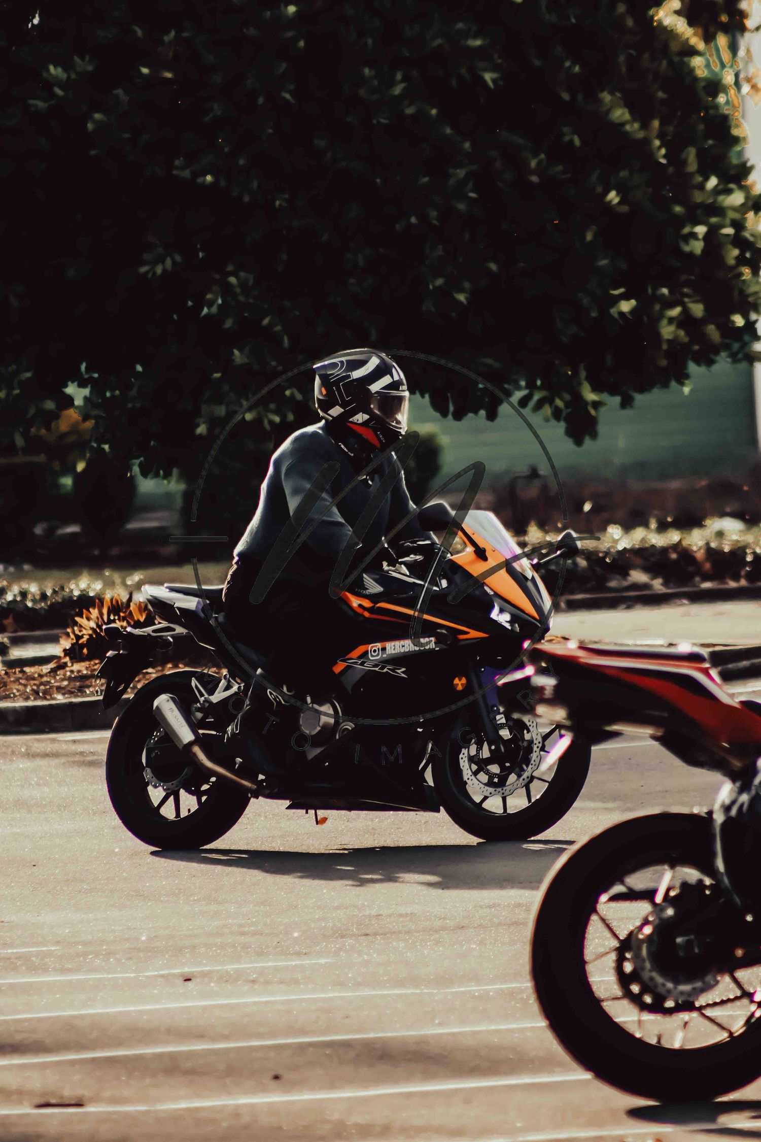 Motorcyclist in black helmet and gear riding an orange KTM sport bike on a sunlit road with trees in background.