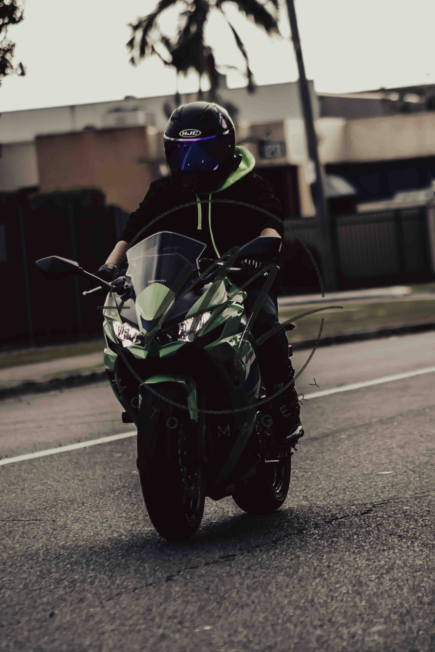 A motorcyclist in a black helmet rides a green Kawasaki sport bike on a city street at dusk.