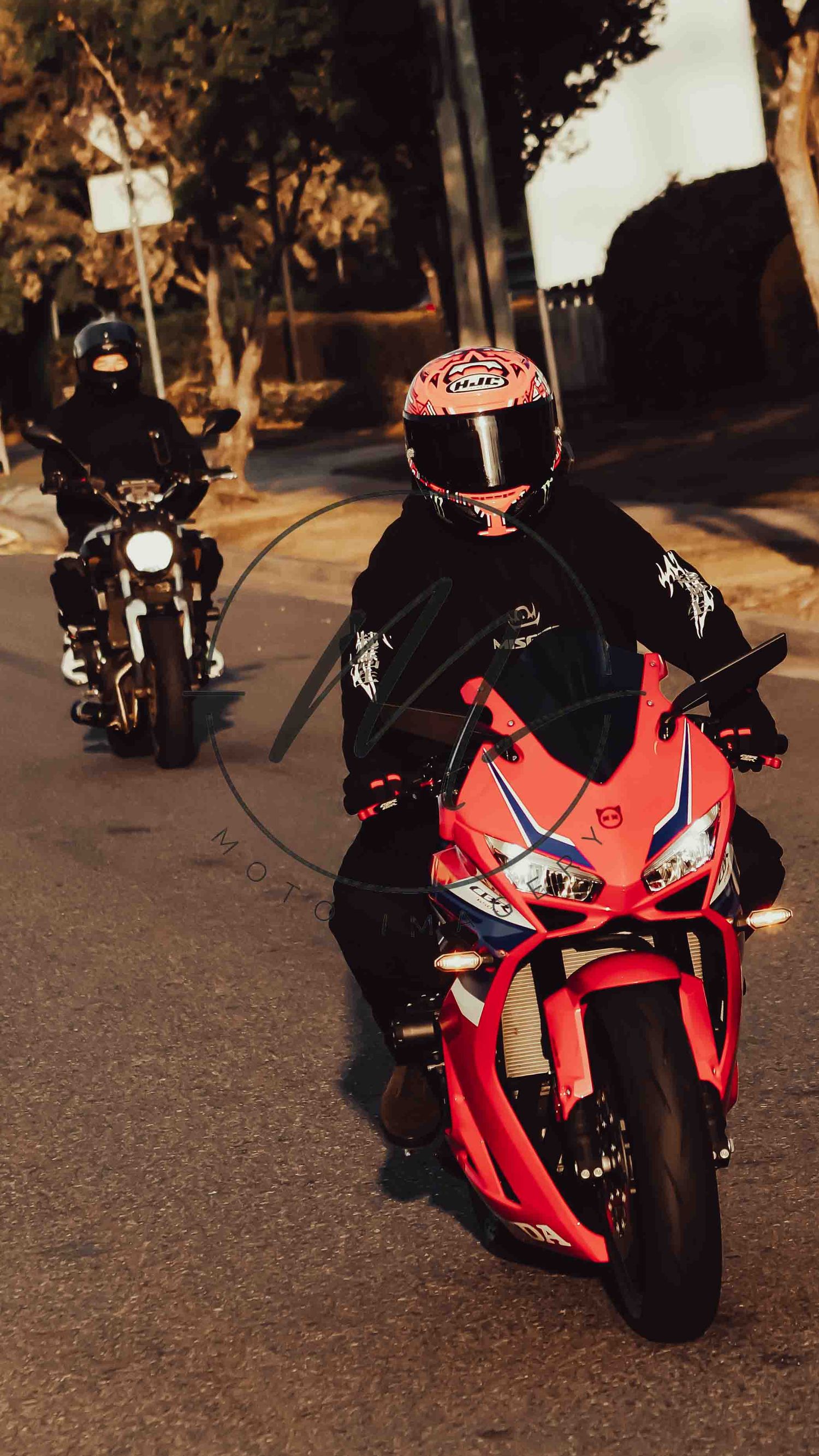 Two motorcyclists riding at night, one on a red sport bike wearing a colorful helmet, another on a dark cruiser behind.