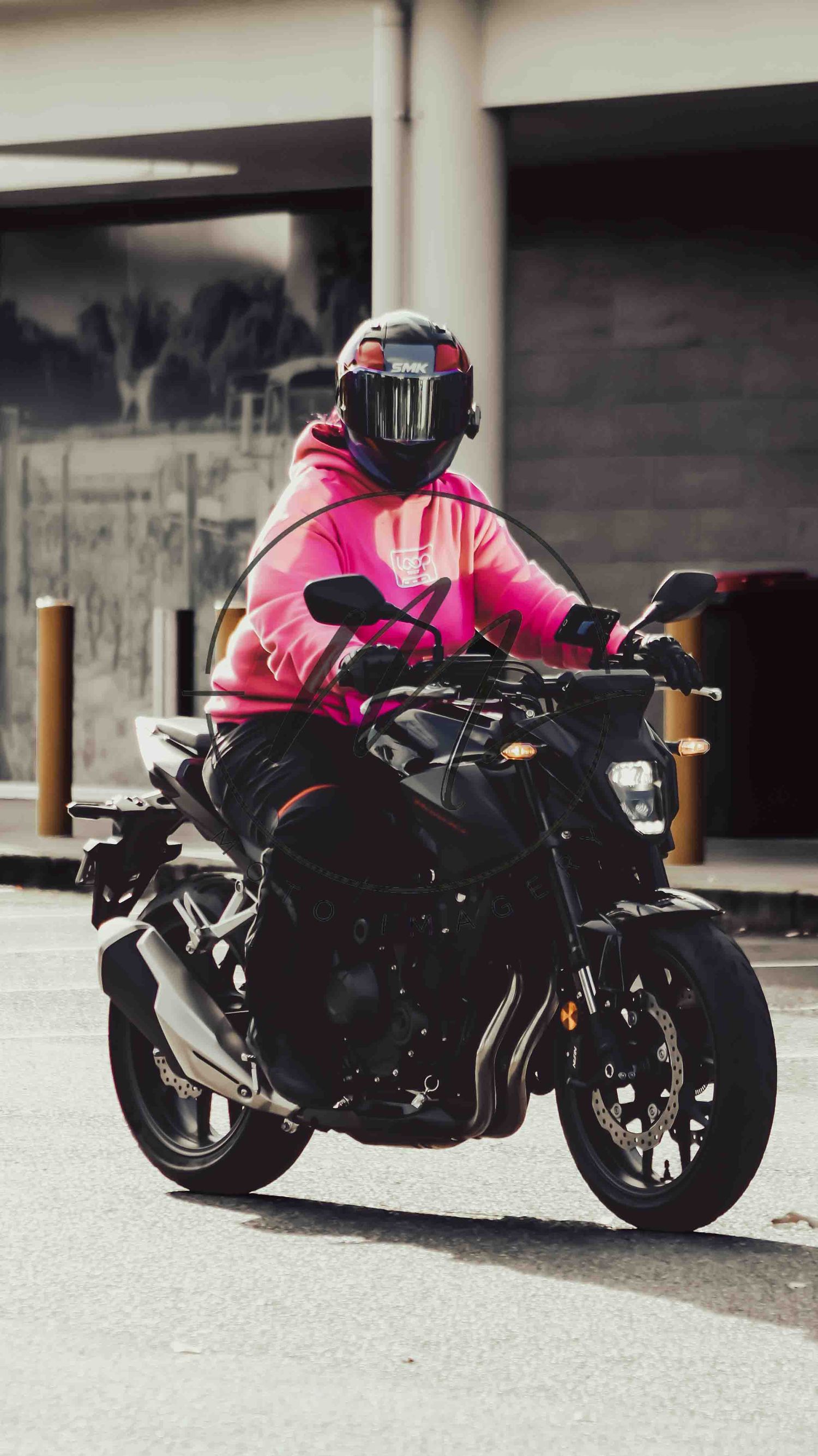 Rider in pink hoodie and black helmet on a dark naked motorcycle on an urban street.