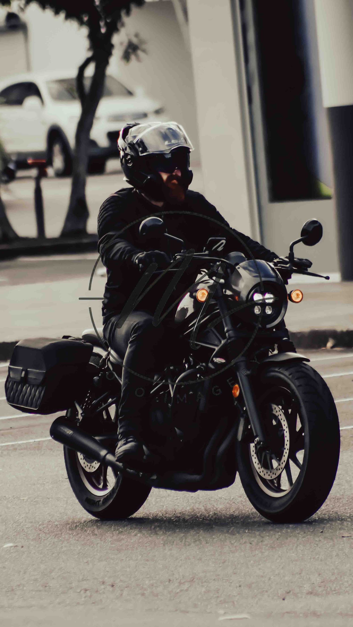 A motorcyclist in all-black leather gear and helmet rides a black cruiser motorcycle on a city street.