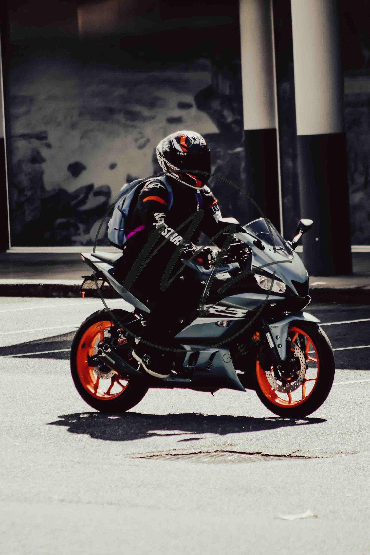 Motorcyclist in full gear riding a black and orange sport motorcycle on a sunny urban street.