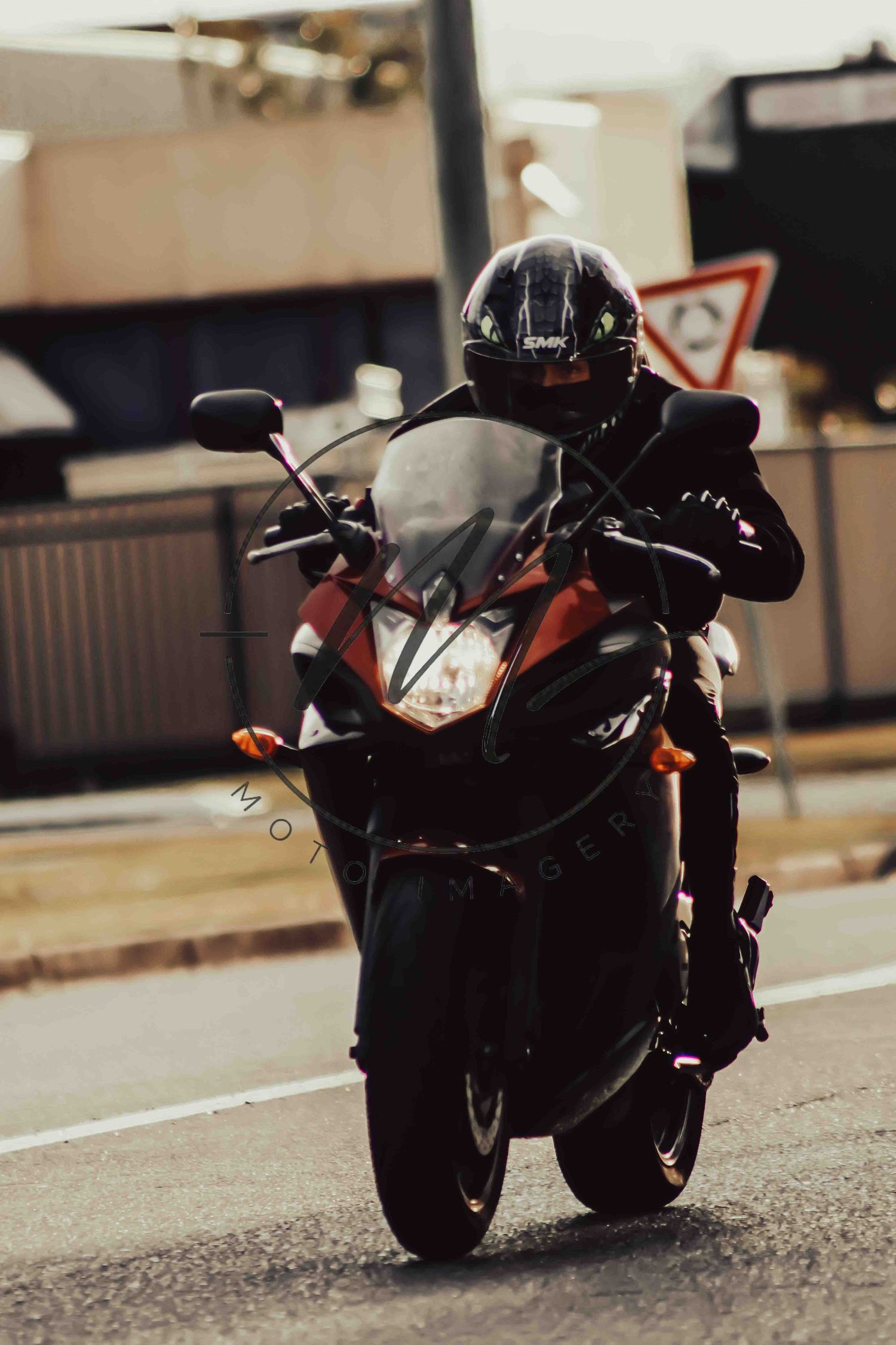 Motorcyclist in black helmet and gear riding a red sport motorcycle on an urban road.