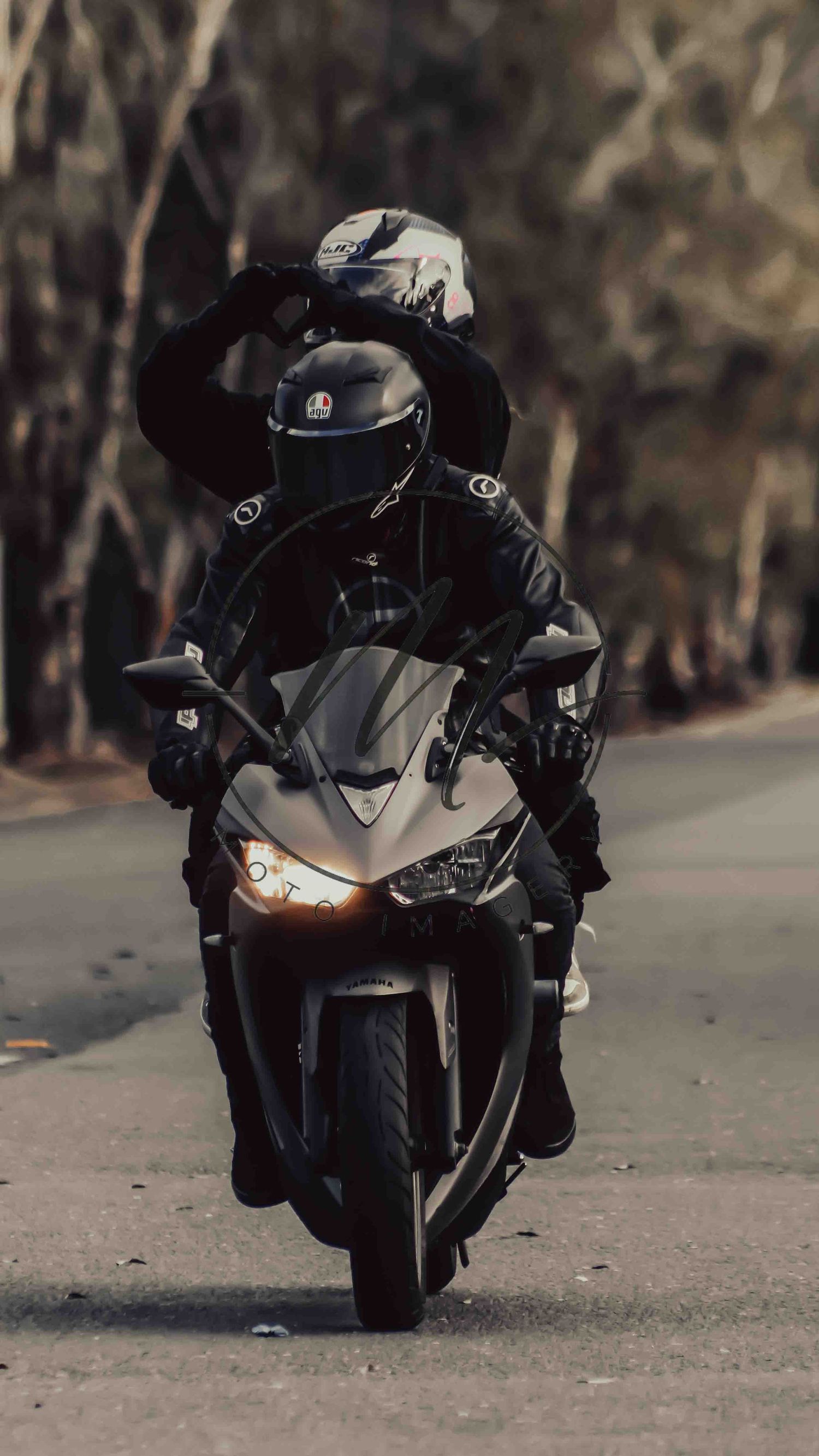 Two motorcyclists in black gear and helmets riding a sport motorcycle on a tree-lined road.