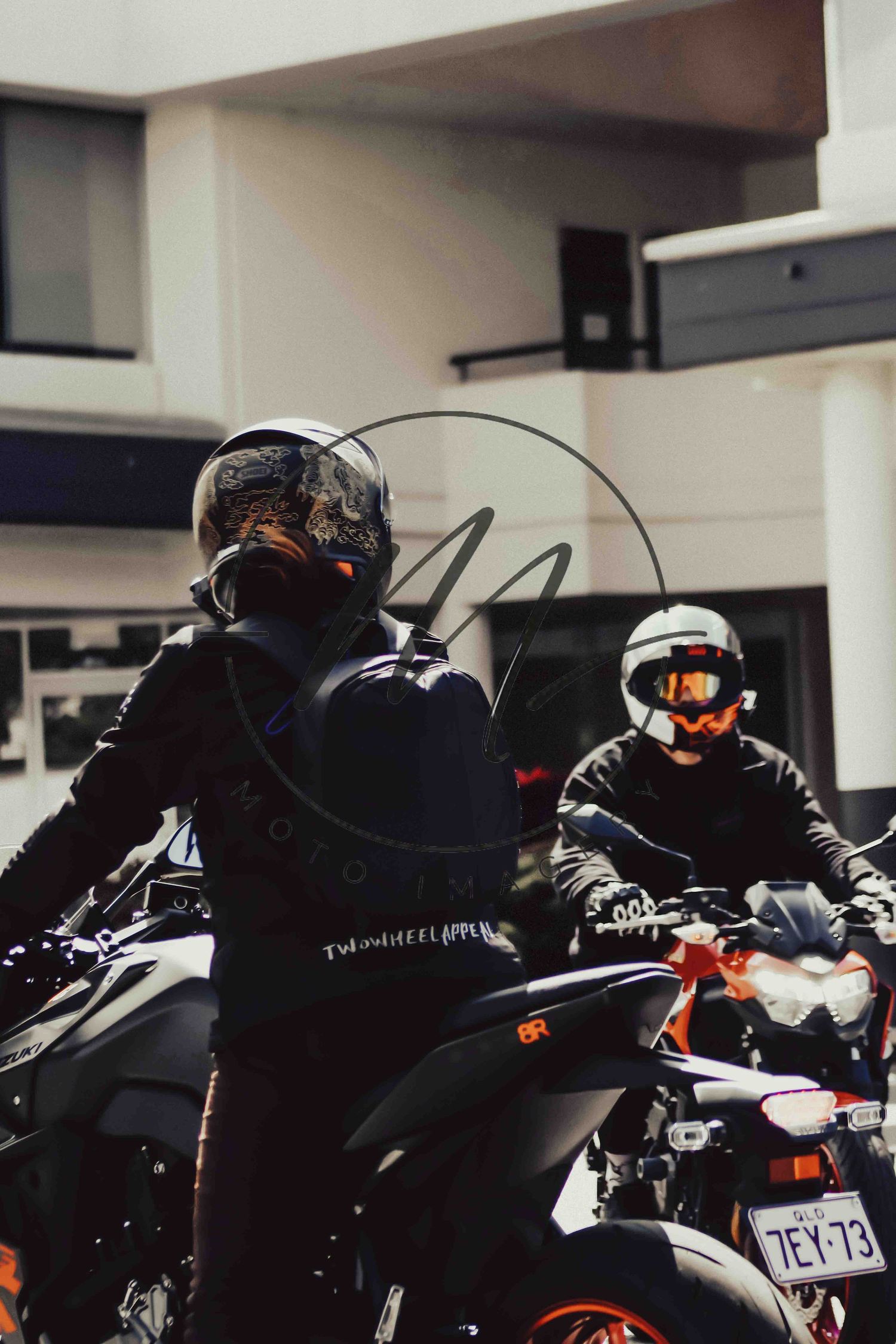 Two motorcyclists in helmets and black gear sit on bikes in an urban parking area, one wearing orange goggles.