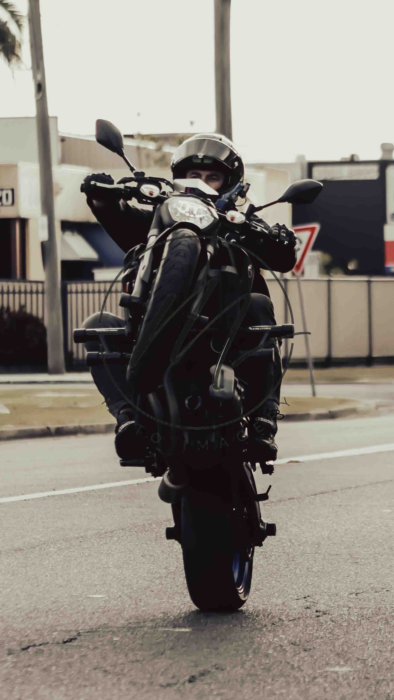 A motorcyclist in full black gear and helmet performs a wheelie on a dark sport bike on a city street.