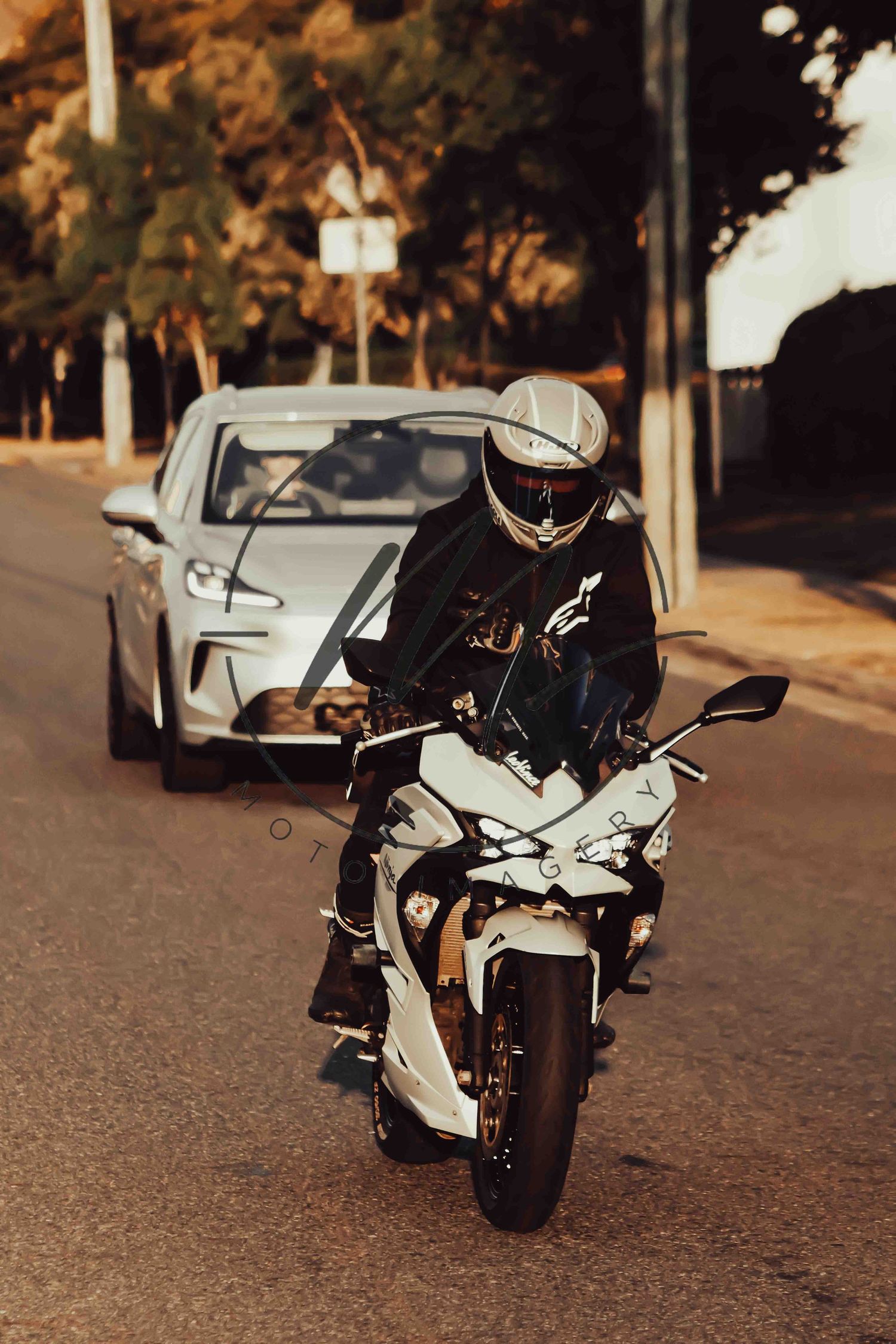 Motorcyclist in black leather jacket and helmet riding a white sport bike on a city street at dusk.