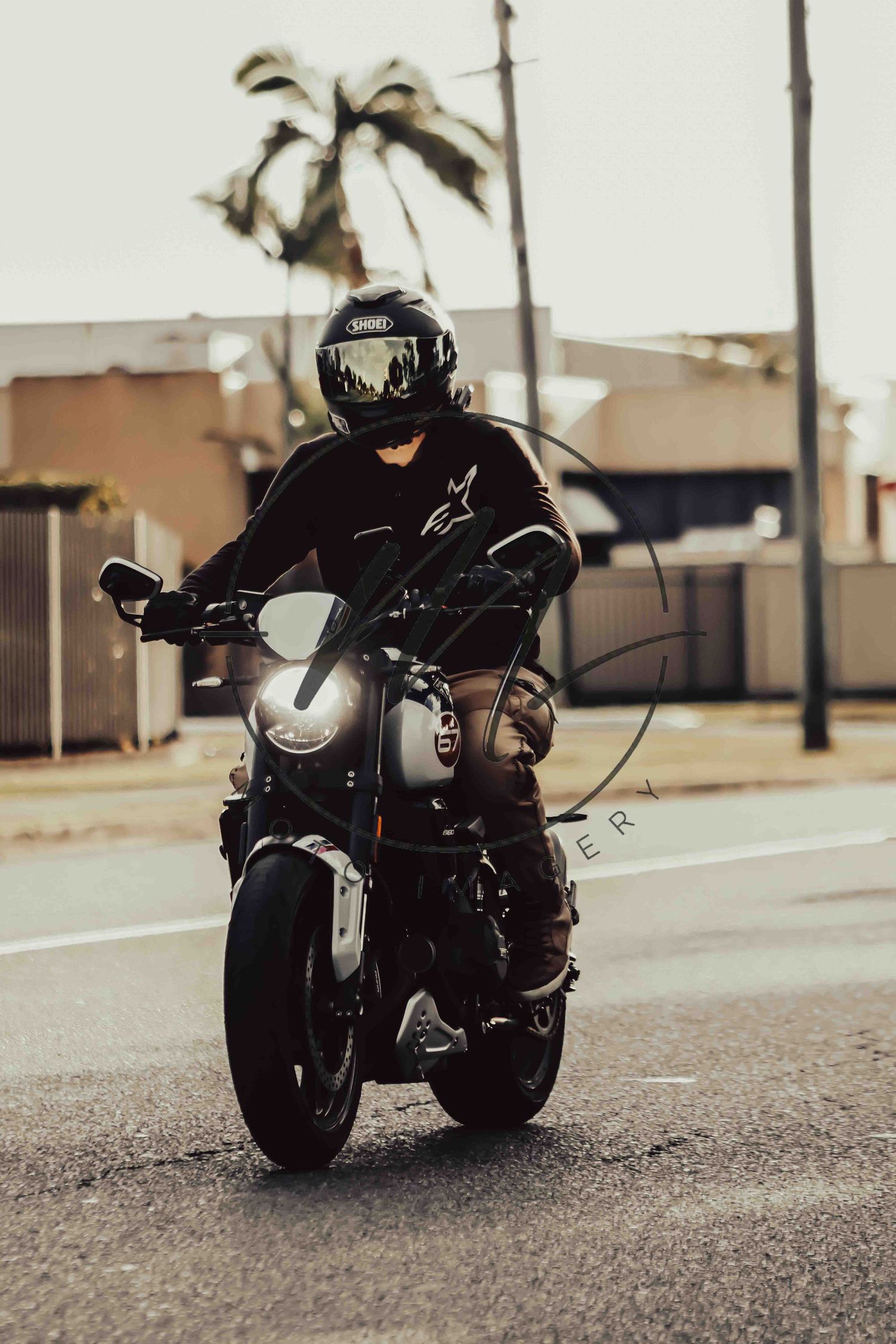 Motorcyclist in black helmet and jacket riding a dark motorcycle on a urban road with palm trees in background.