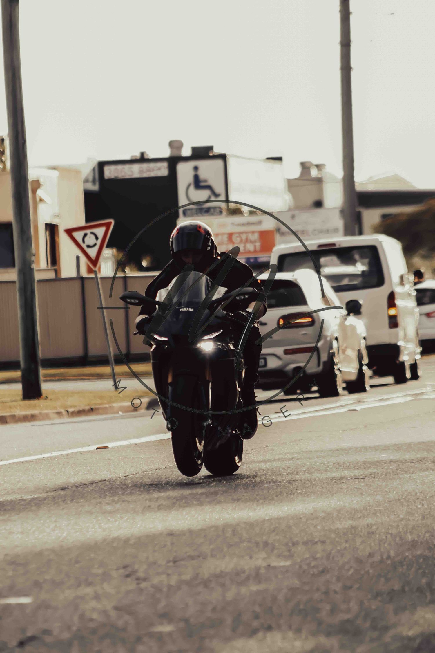 A motorcyclist in black gear rides on a busy urban road with cars and a disability sign visible in the background.