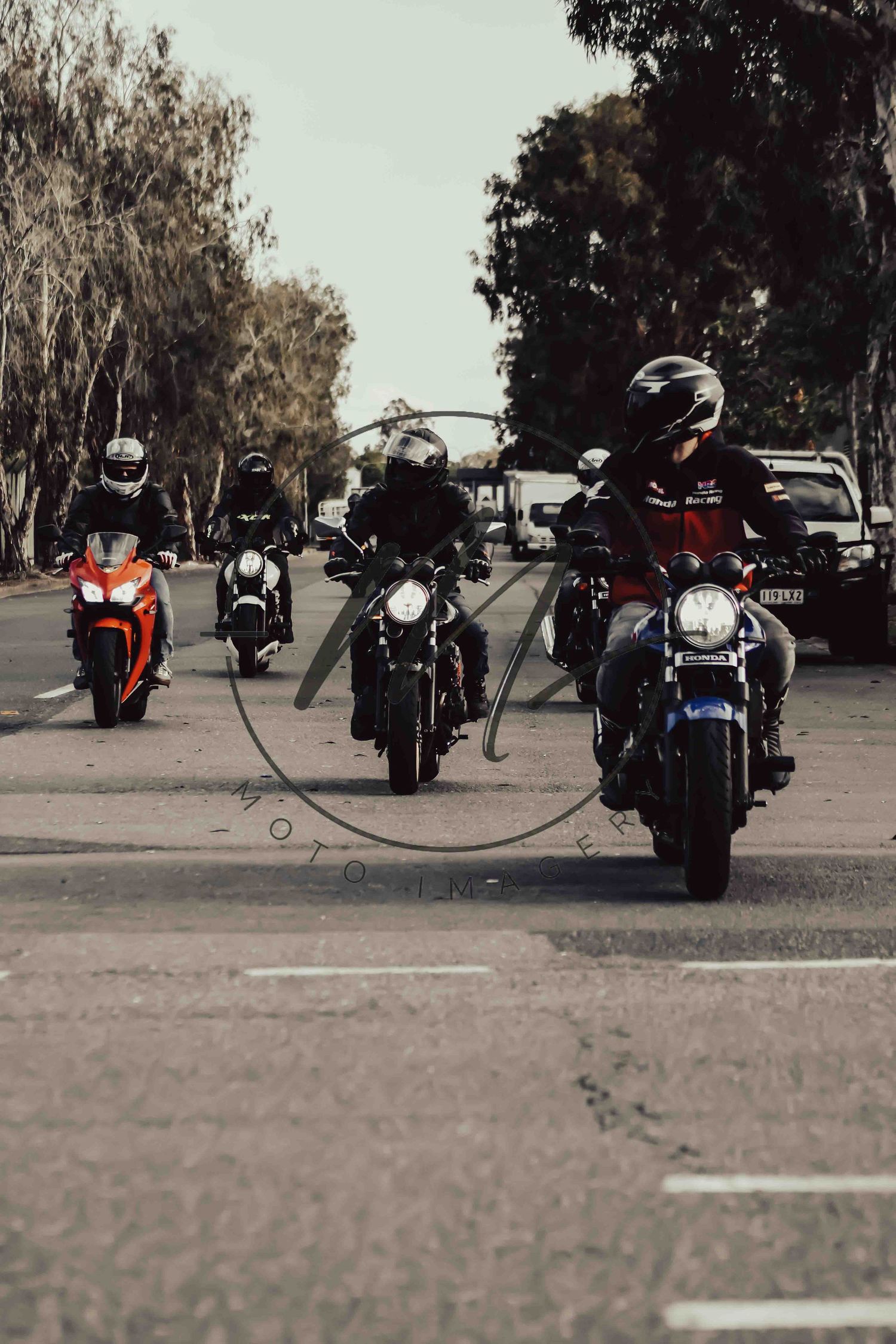 Group of motorcyclists riding in formation down a tree-lined road, wearing helmets, leading with a blue bike.