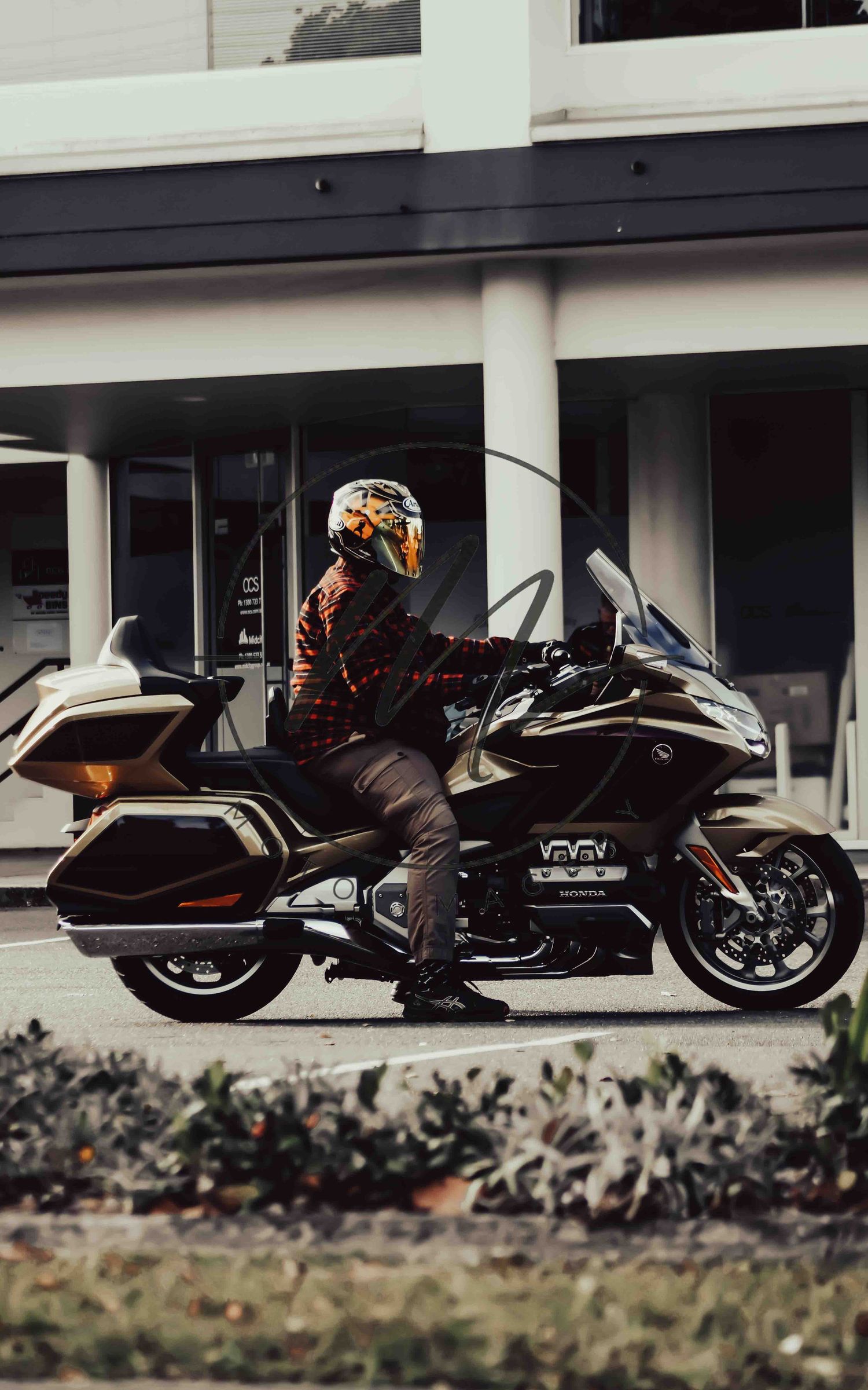 Rider in a plaid jacket and white helmet sitting on a Honda Gold Wing touring motorcycle parked outside a modern building.
