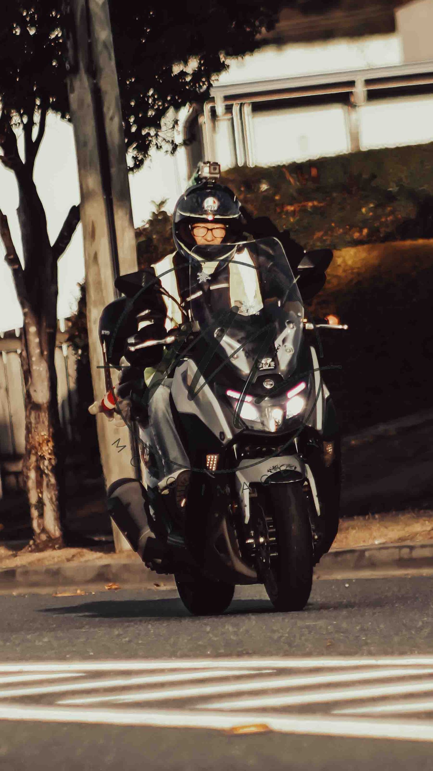 A motorcyclist in a helmet rides a modern scooter on a city street lined with trees and buildings.