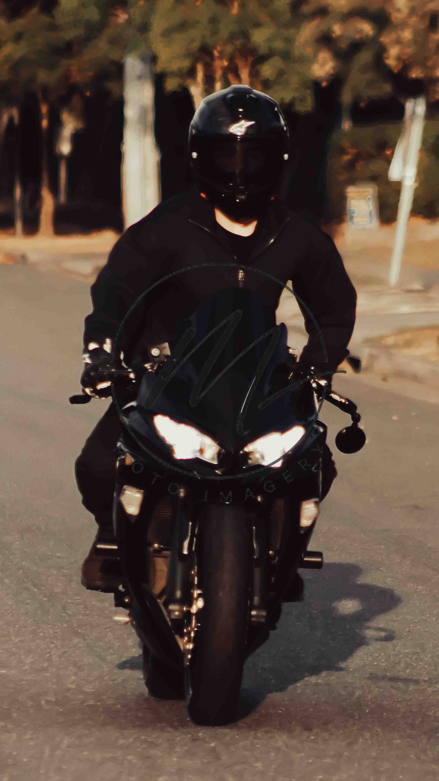 A motorcyclist in all-black gear and helmet rides a black sport motorcycle on a road at dusk.