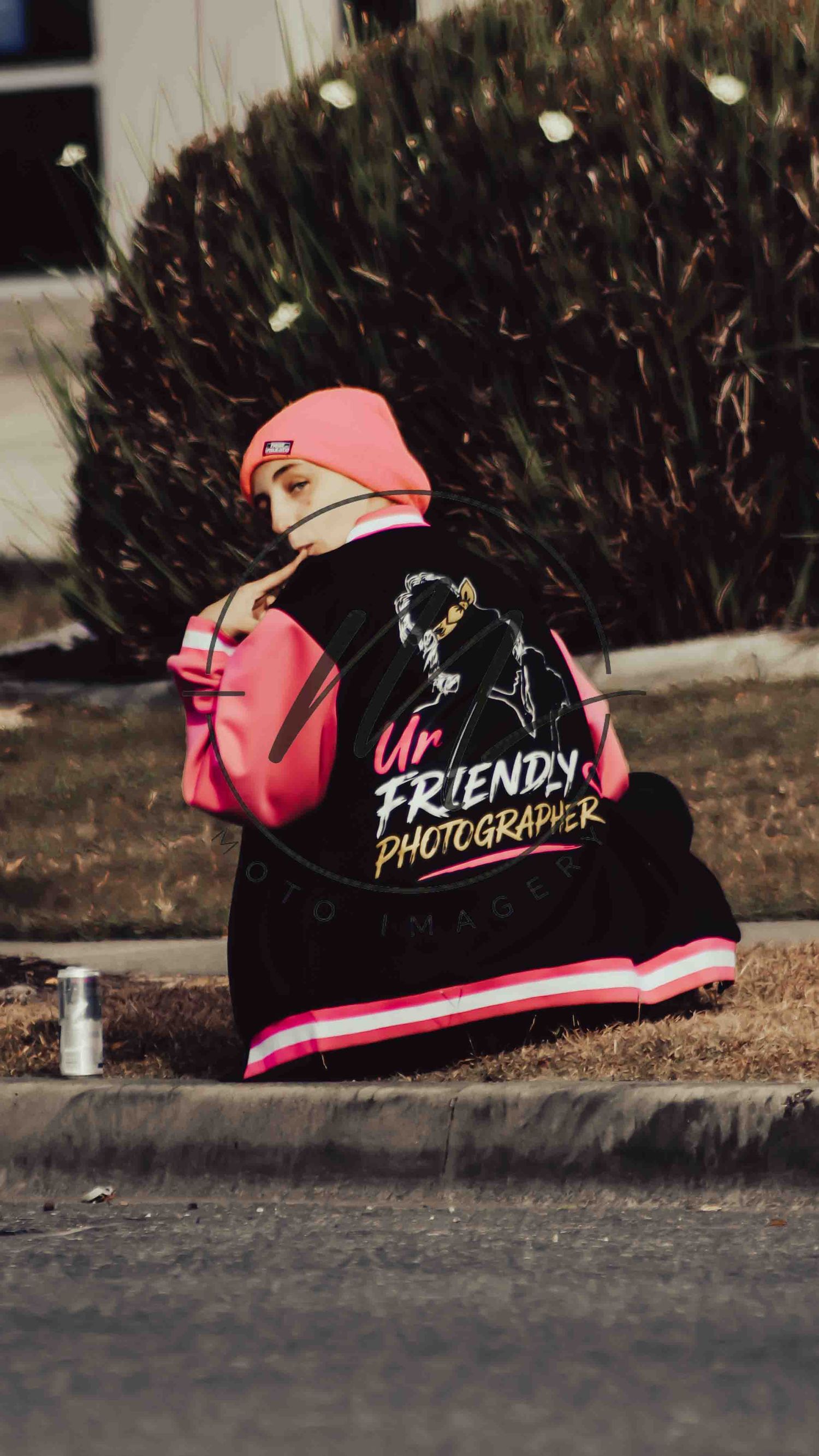 Person sitting outdoors wearing a black varsity jacket reading 'Ur Friendly Photographer' with pink accents and beanie.