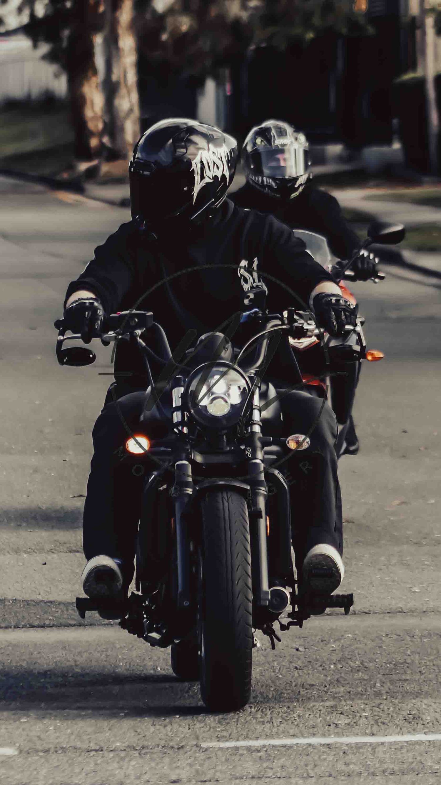 Two motorcyclists in black helmets and jackets ride motorcycles down a street in a moody urban setting.