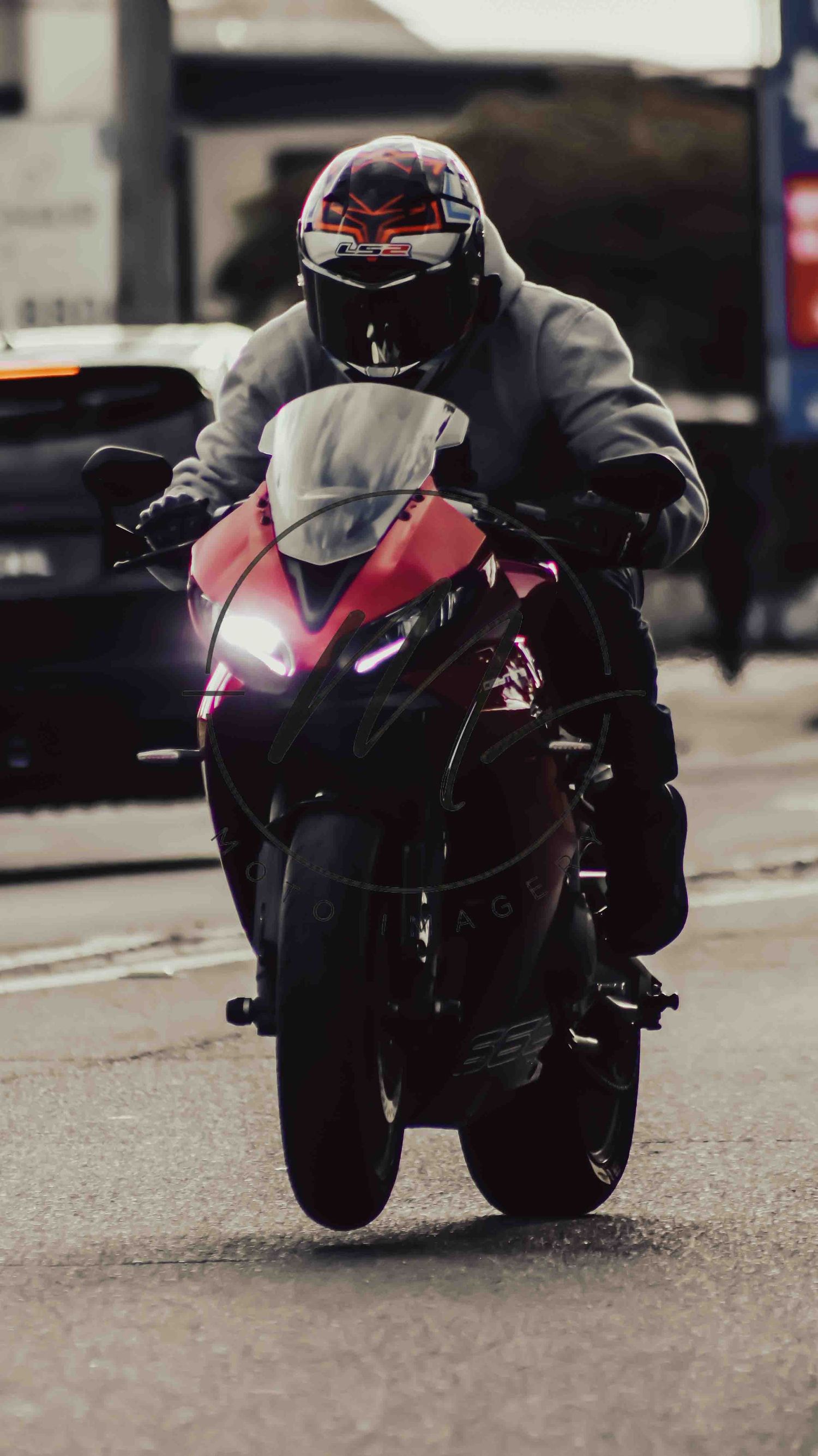Rider on red sport motorcycle wearing gray hoodie and decorated helmet performs wheelie on urban street.
