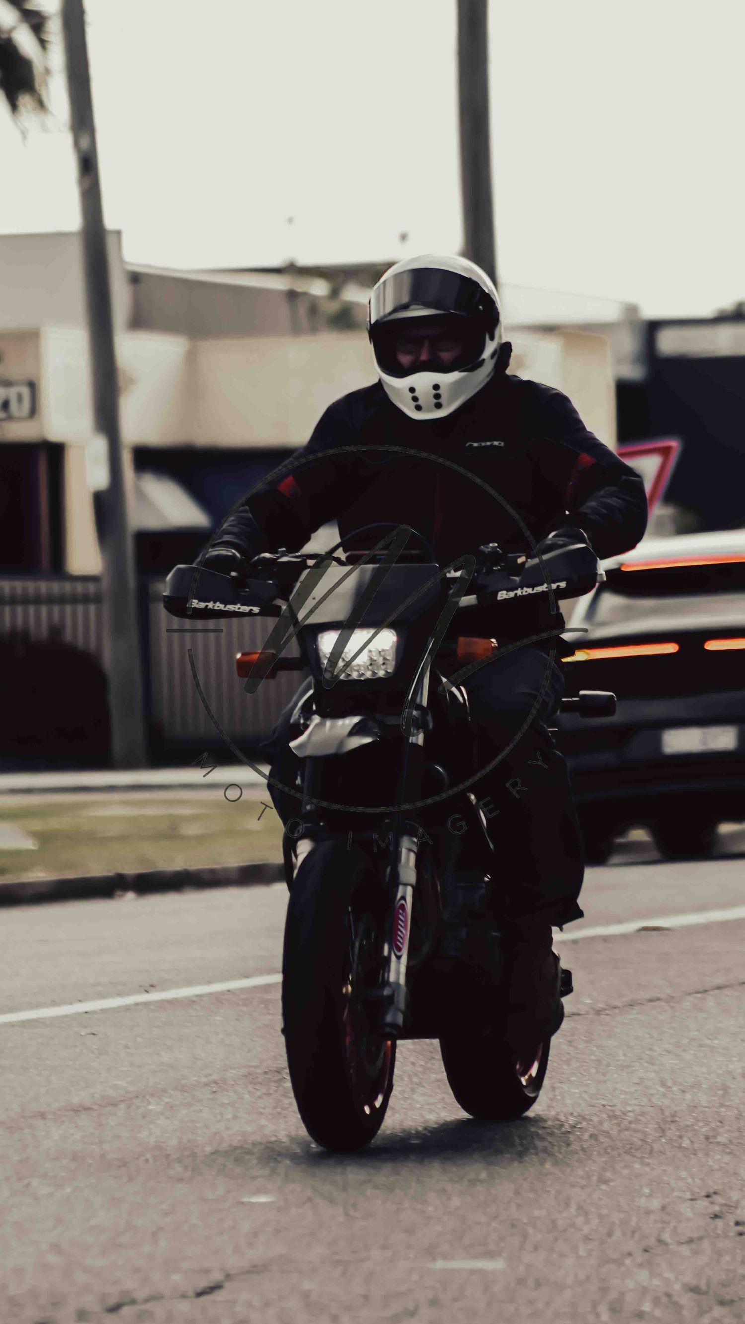 Motorcyclist in black gear and white helmet riding a dark sport bike on a city street.