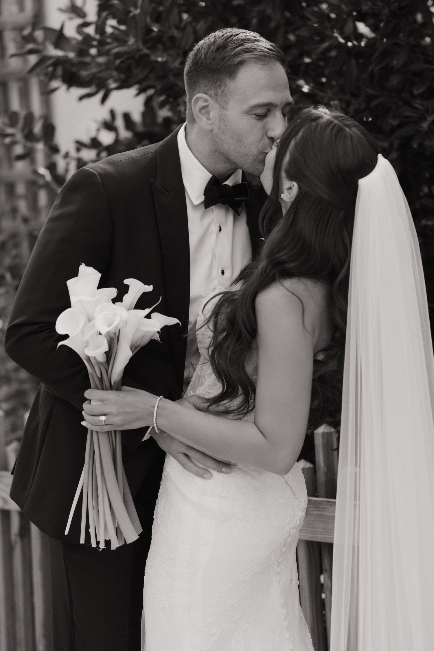 Black and white photo of a bride and groom sharing a kiss, bride holding calla lily bouquet with veil flowing.