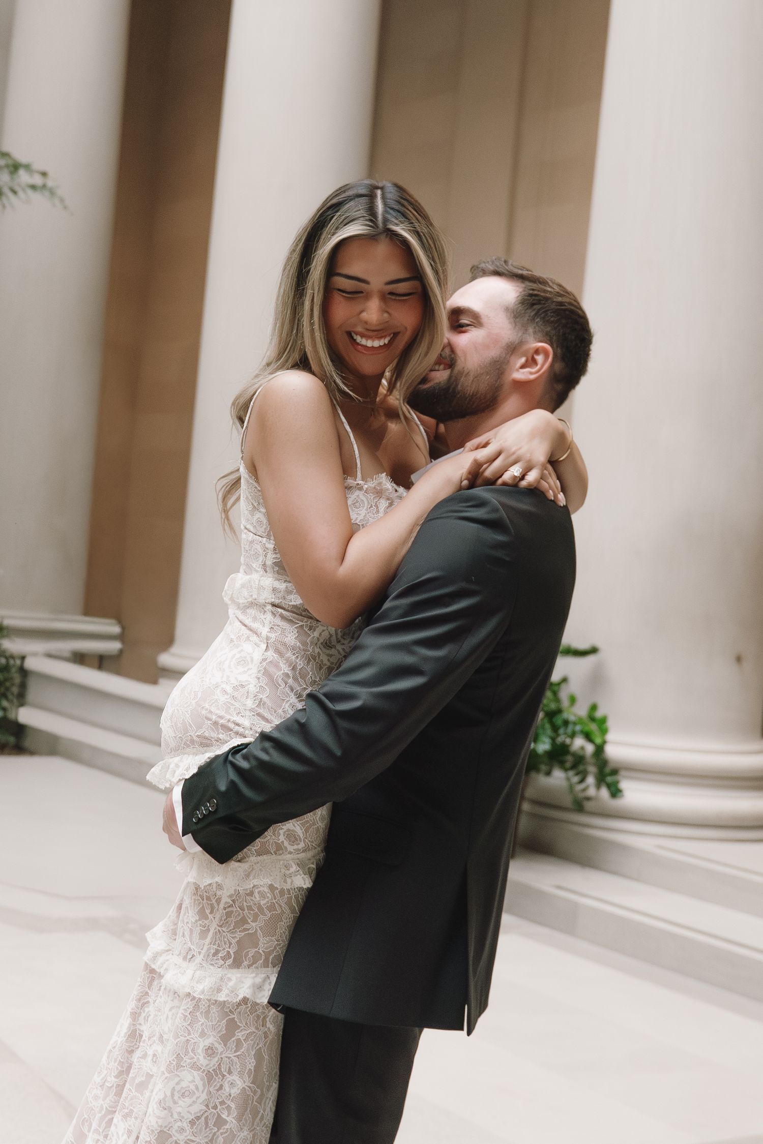 Joyful bride in lace gown laughing as groom in black suit kisses her cheek by columns.