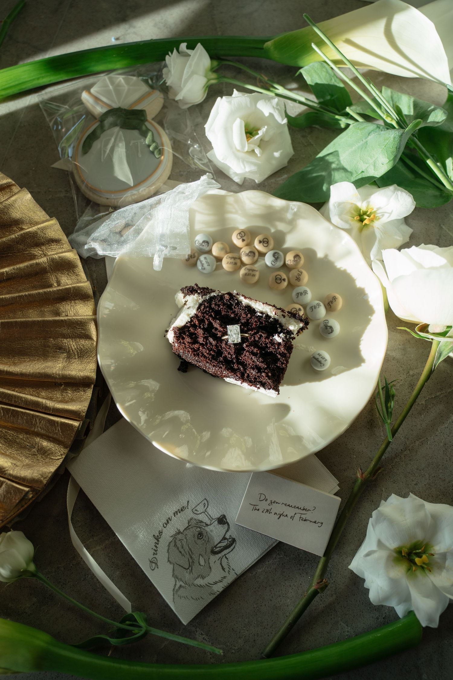 Overhead flat lay of chocolate cake slice, white flowers, and proposal note on table.