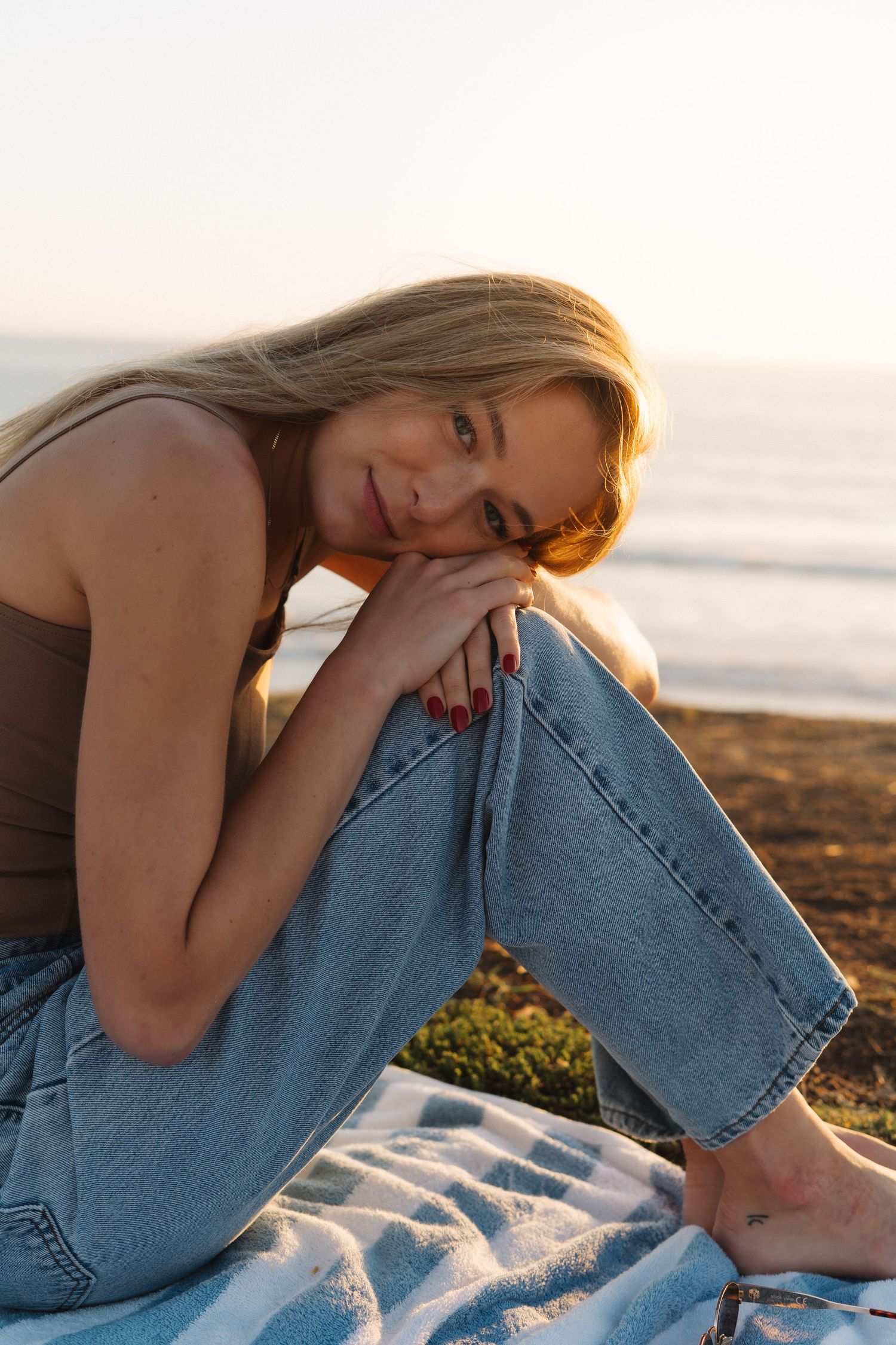 Blonde woman sitting on a blanket at the beach during golden hour, smiling at the camera.