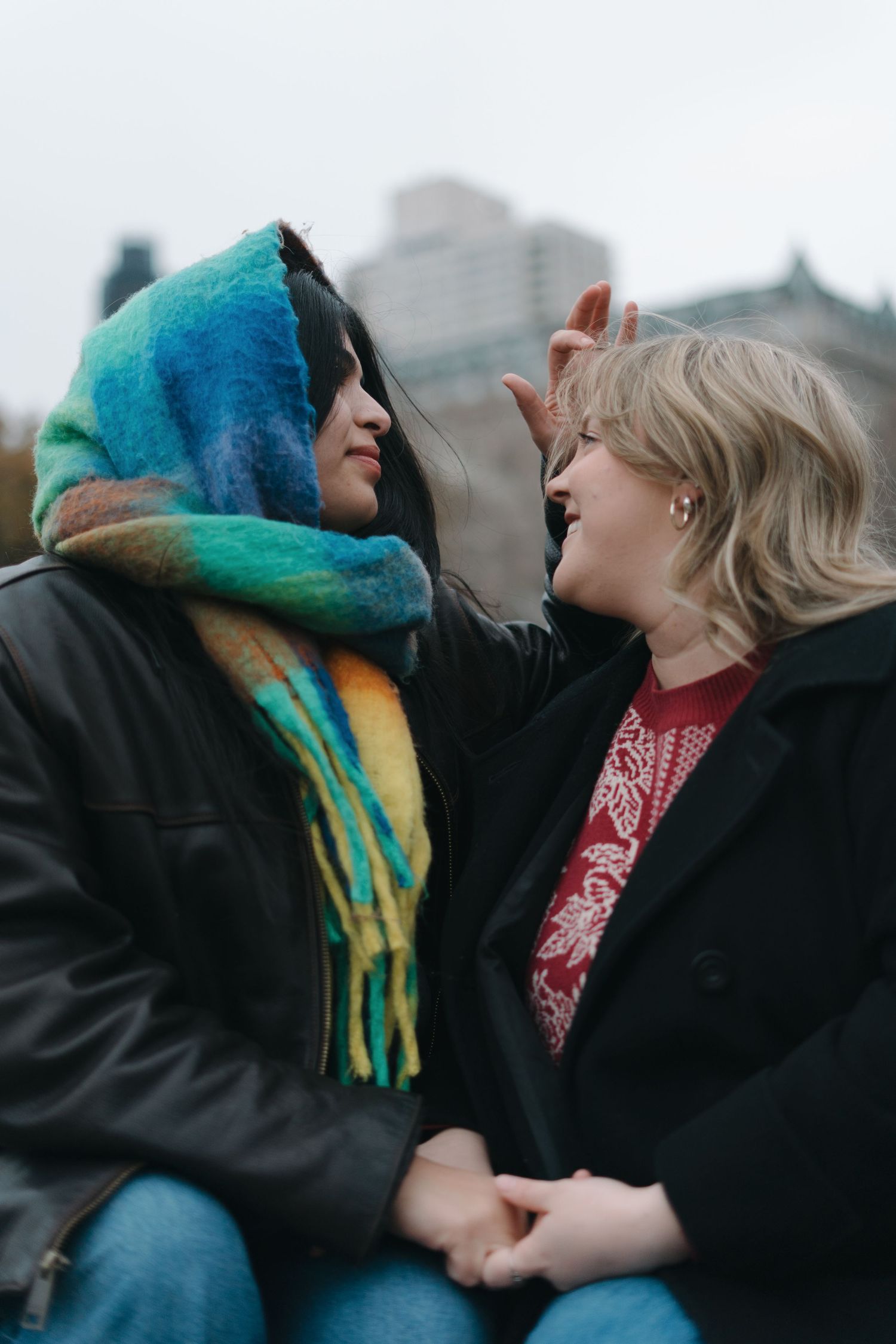 Two women laughing and holding hands outdoors, one wearing a colorful rainbow scarf.